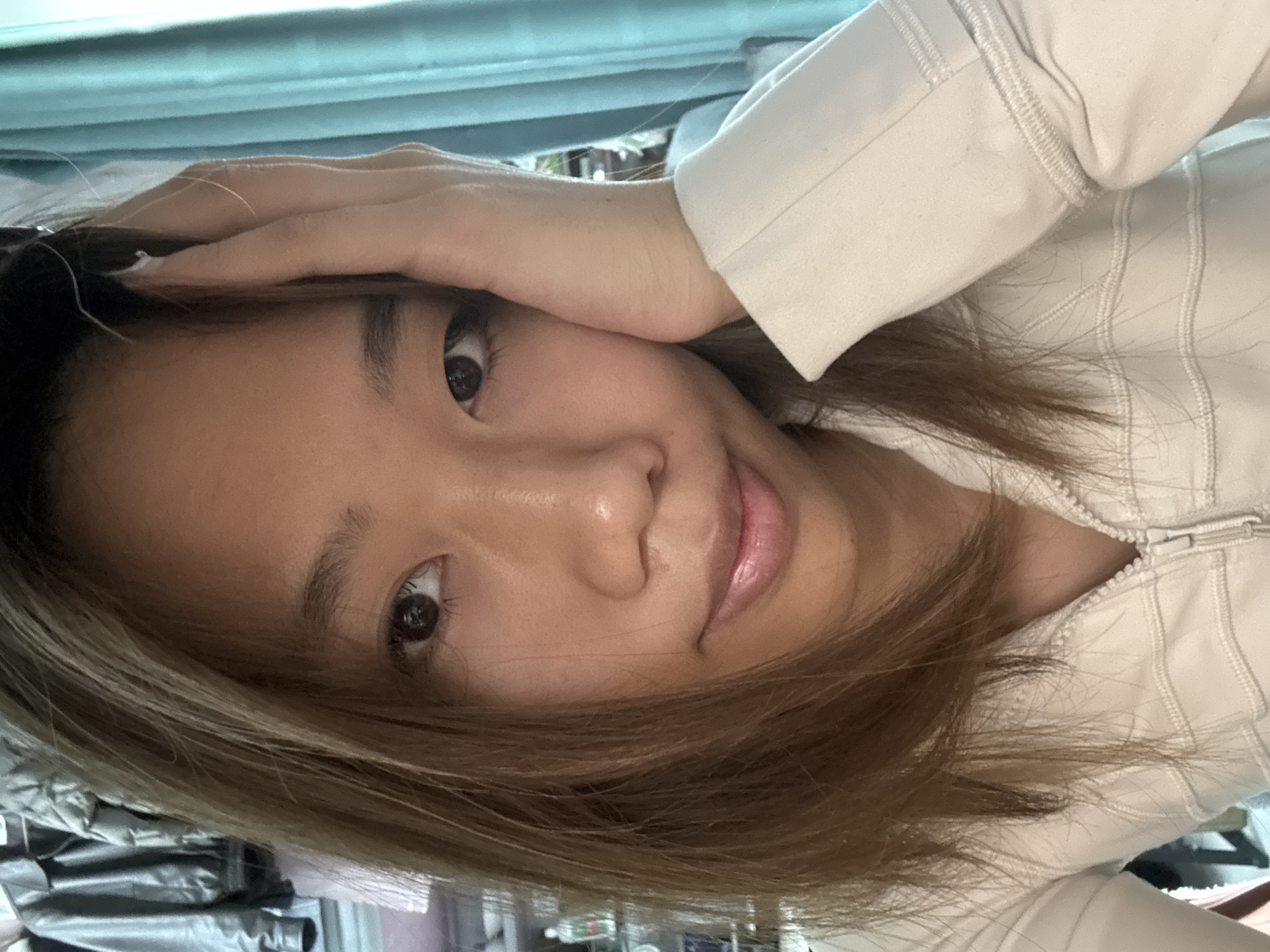 A close-up, slightly angled portrait shows a young woman with light brown hair partially obscuring her face. Her right hand is raised, with fingers near her forehead. She has brown eyes and appears to be smiling slightly. She is wearing a white zip-up jacket. The background is blurred and shows a hint of daylight through a window. There is no indication of Nan'an District, China.