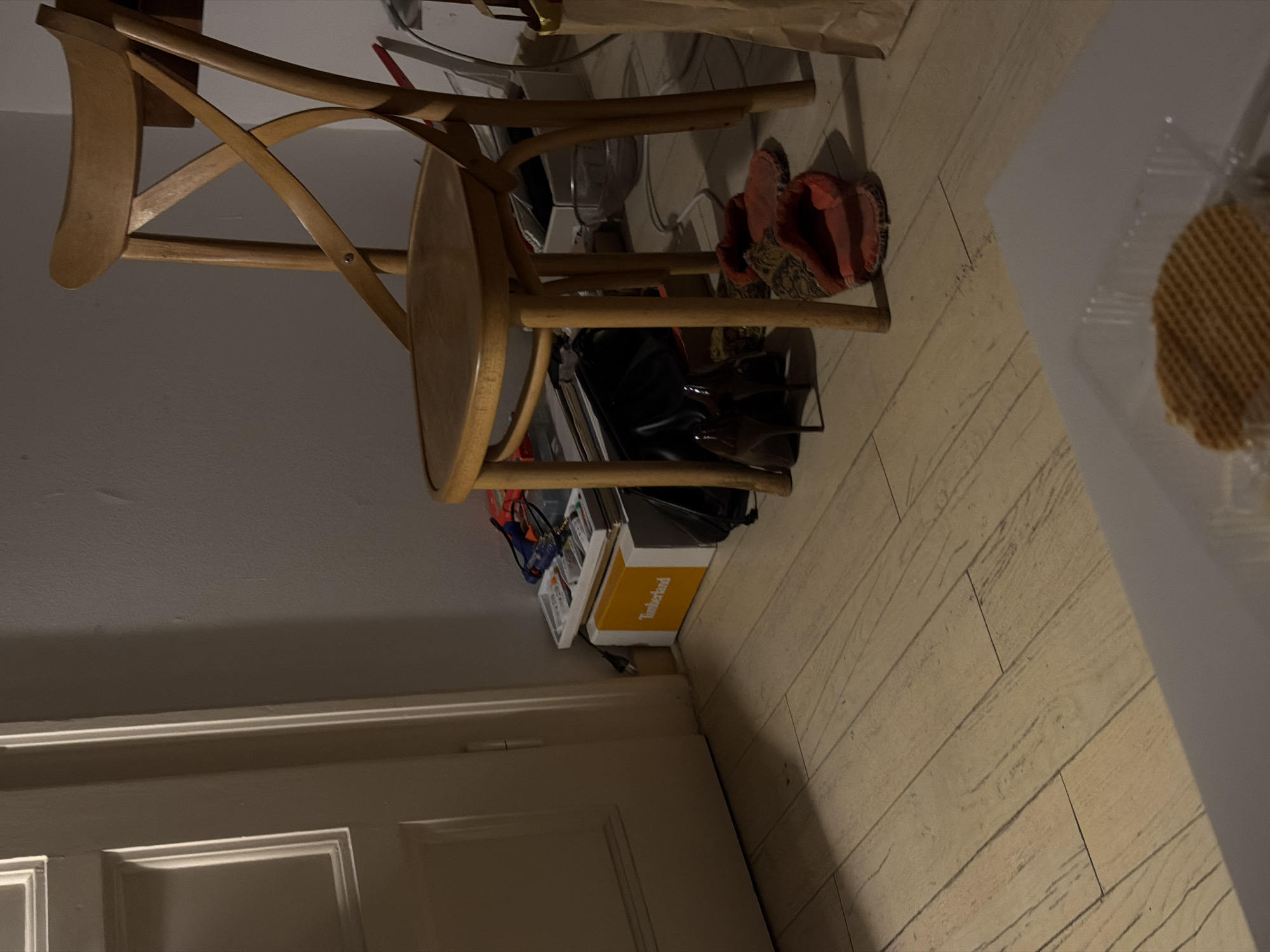 An interior shot, likely a domestic setting, in the Vracar Urban Municipality, Serbia, captures a dimly lit room with a wooden floor. A light-colored door is partially visible on the left. In the center, a wooden chair with a cross-back design stands. Beneath the chair, a pile of items includes a Timberland shoebox, a black bag, a pair of brown high-heeled shoes, and a pair of red slippers. To the right of the chair, a portion of a paper shopping bag is visible. The scene is illuminated by low lighting, casting shadows.
