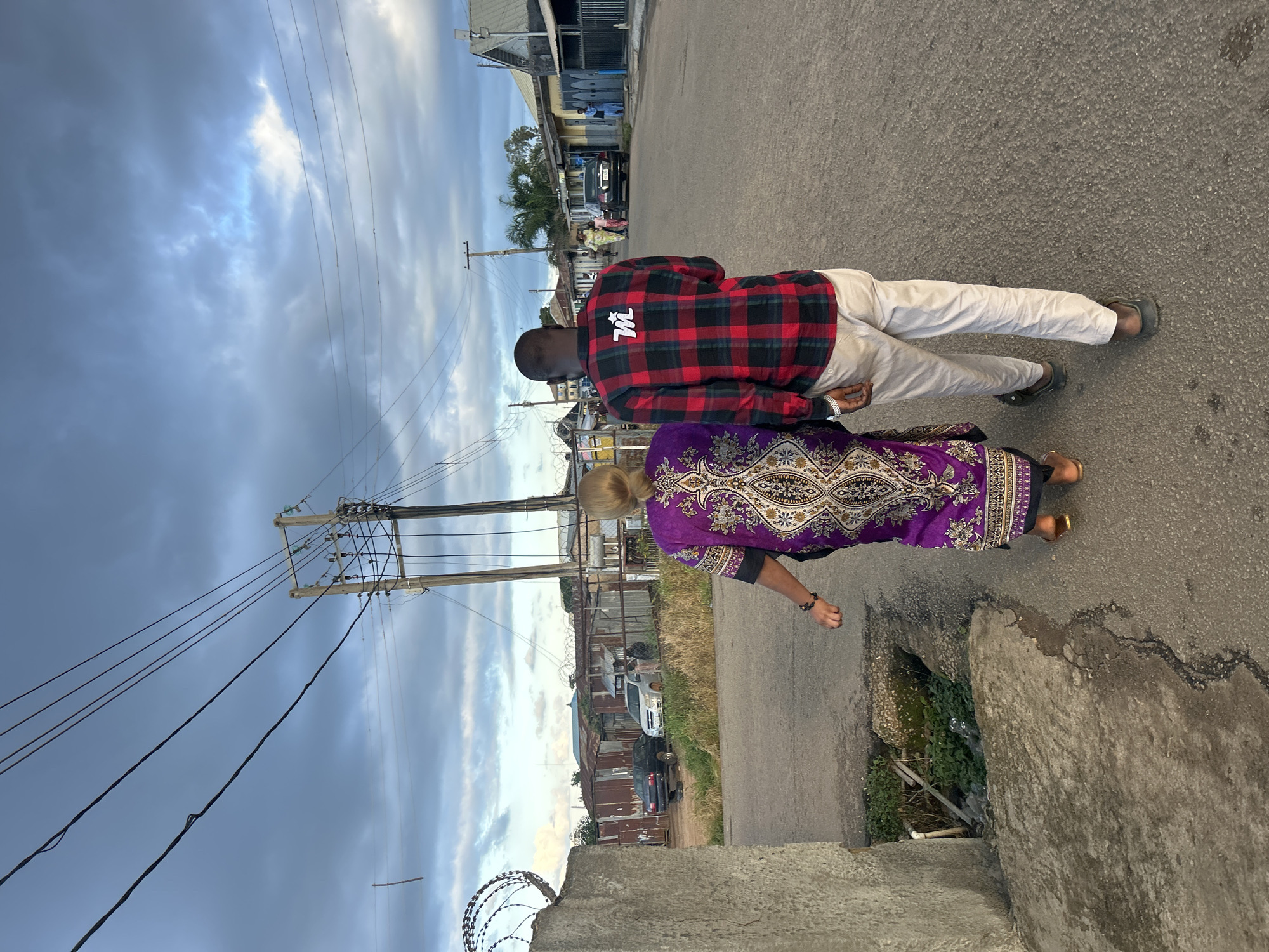 An unidentifiable person with blond hair, from the back, wearing a long, purple, patterned dress and brown sandals, walks on an asphalt road. They are accompanied by an unidentifiable person with dark skin, from the back, wearing a red and black checkered long-sleeved shirt with a white logo on the back and light-colored pants with black sandals. They are walking away from the camera and toward other people and buildings in the distance. The sky is cloudy and appears to be twilight. The location is identified as Jos, Nigeria.
