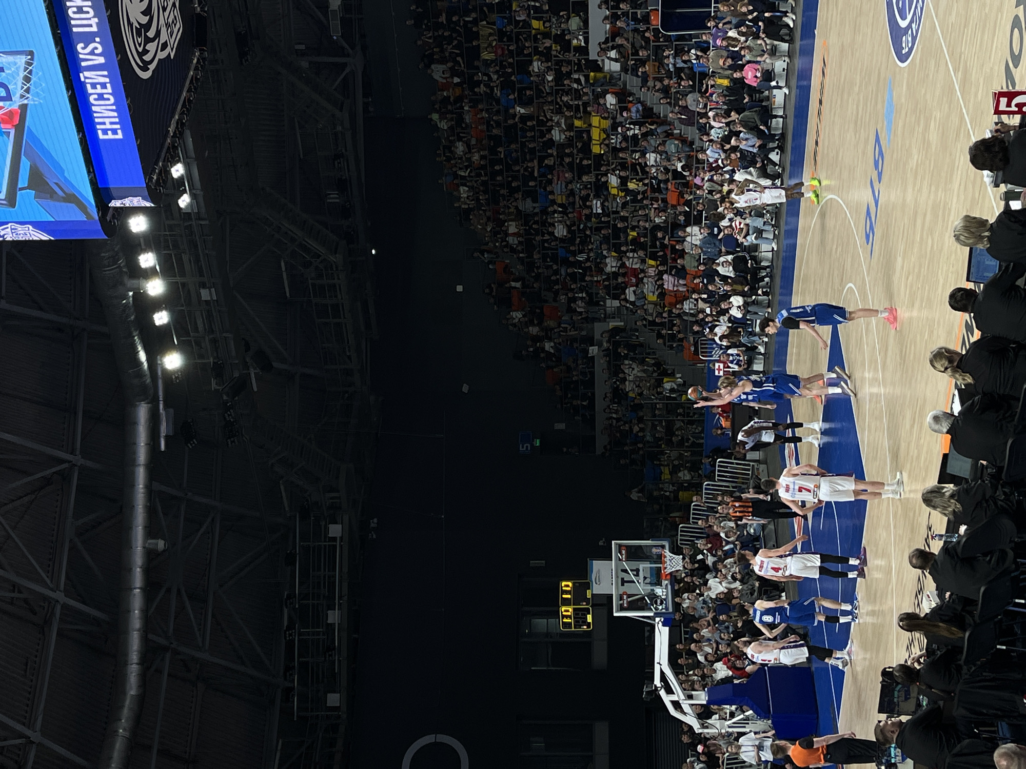 The image shows a basketball game in progress at the Arena Sever in Krasnoyarsk, Russia. Players in blue and white uniforms are positioned on the court near the basket, with one player in blue leaping to shoot the ball. The scoreboard shows the game clock at 10:10. Spectators fill the stands, and staff members are seated along the sidelines. The arena's interior reveals exposed structural elements and lighting fixtures. The overhead screen displays the game matchup in Russian.