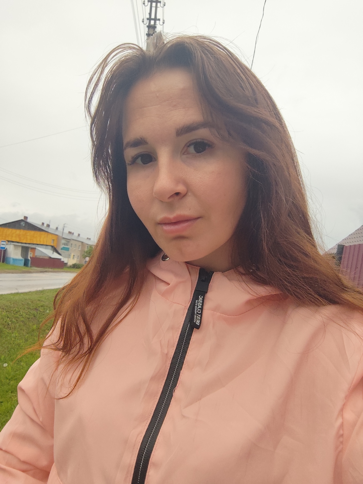 A woman is depicted in a headshot selfie, looking directly into the camera with a neutral expression. She has long, wavy, reddish-brown hair that frames her face and flows over her shoulders. She is wearing a light pink hooded jacket featuring a black zipper down the front. A black zipper pull displays white text, which appears mirrored but reads "BEST CLASSIC" and "STAY STRONG WINS ALL".
The setting is outdoors, under an overcast, bright sky, suggesting a cloudy daytime. In the background, what appears to be a street scene in Фоки, Russia, unfolds. On the left, a multi-story building with a distinctive yellow upper facade and brown roof stands out, with other residential-style buildings stretching into the distance. A road or paved area is visible, bordered by a strip of green grass. A utility pole and wires are present towards the top center. On the right side, a portion of a reddish-brown fence or roof is visible. The overall atmosphere is calm and everyday.