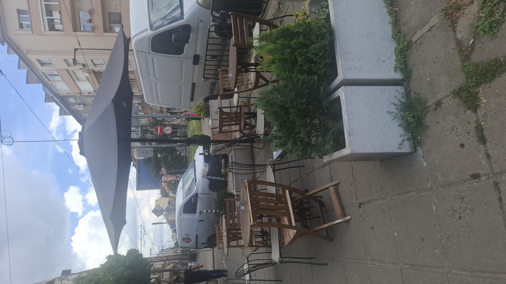 The image shows an outdoor seating area for a cafe on a city sidewalk in Sofia, Bulgaria. Several wooden tables and chairs are arranged under a large, dark umbrella. A white delivery van is parked on the street behind the tables, and a large white van is parked further back. Buildings line the street, and there are power lines and street lights overhead. The sidewalk is paved with concrete tiles. Two concrete planters with small evergreen trees are positioned in front of the seating area. The sky is cloudy.