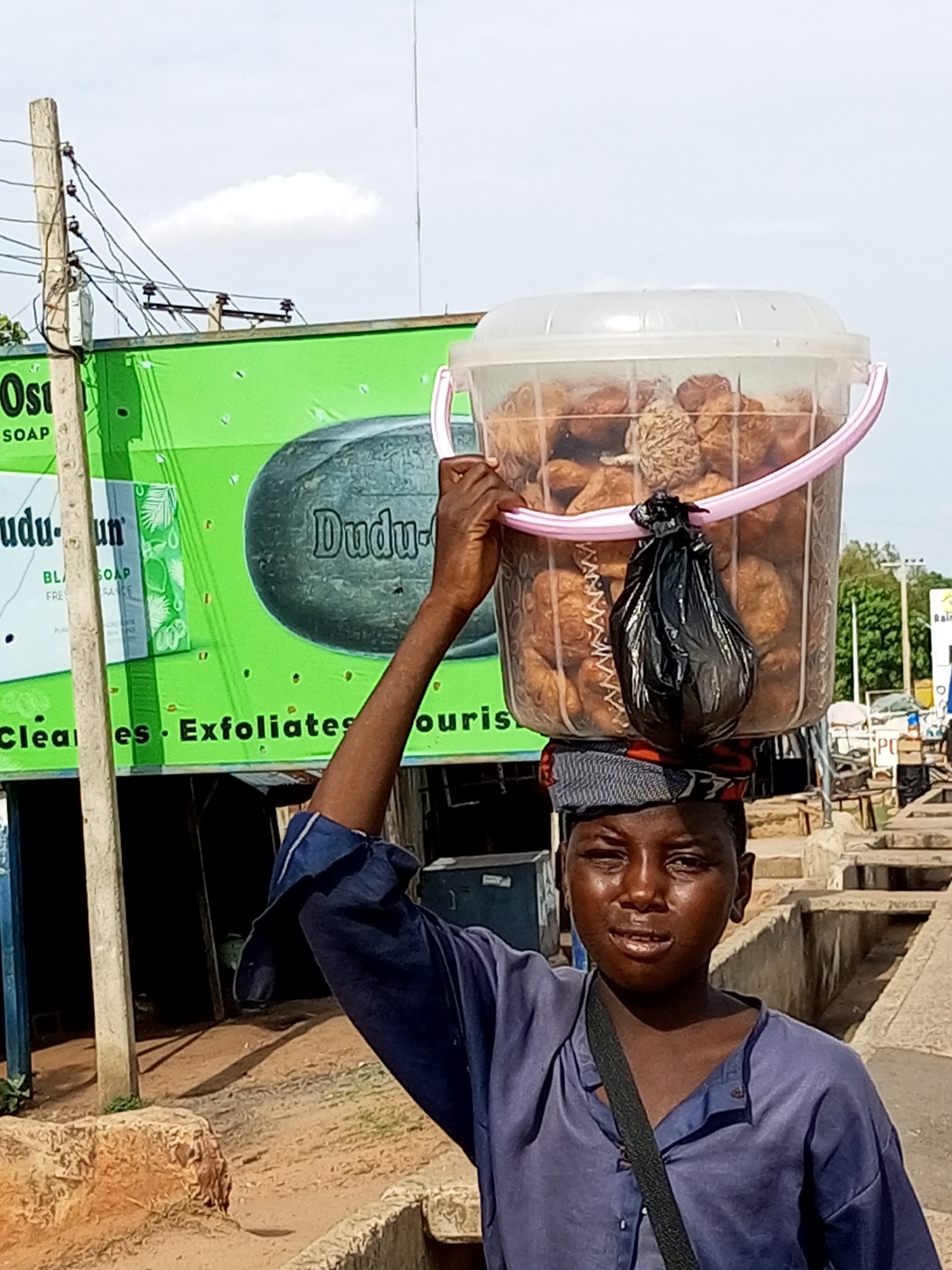 A young person, likely a vendor, carries a large, clear plastic bucket filled with food items on their head. The food appears to be small, round, and brown, possibly fried dough or snacks. A black plastic bag is tied to the inside of the bucket, obscuring some of the contents. The vendor wears a blue tunic and a patterned headwrap. They are walking on a dirt path next to a concrete drainage channel. In the background, a bright green billboard for "Dudu-Un Soap" is visible, along with utility poles and wires. The scene suggests an outdoor market or street setting in the city of Gombe, Nigeria, under a cloudy sky. The overall impression is one of daily commerce and street life.