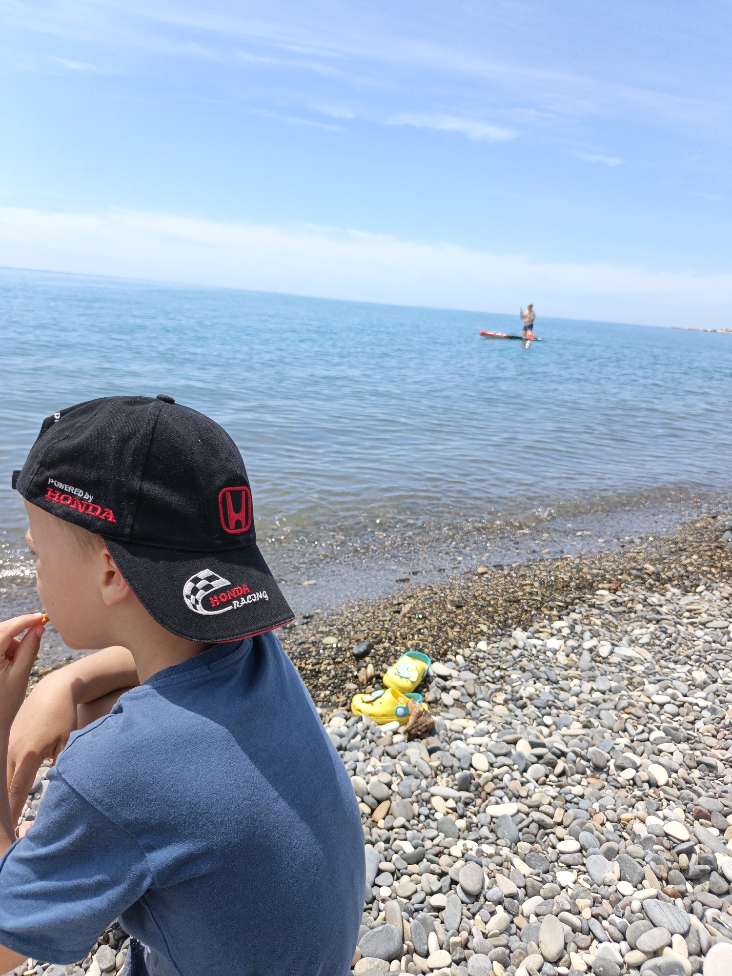 The image presents a sunlit pebble beach scene in Krinitsa, Russia, along the Black Sea coast. In the immediate foreground, positioned to the lower left and center, a young male child is observed from his left profile and back, seated on the multi-colored pebbles. He wears a dark blue short-sleeved t-shirt and a black baseball cap. The cap features "POWERED by HONDA" embroidered in red on the front-left panel, a red Honda "H" logo on the side, and "HONDA RACING" with a checkered flag design on the brim. His left hand is raised to his mouth, holding a small, light-colored food item, possibly a nut or cracker.
The beach itself comprises a dense expanse of smooth, rounded pebbles ranging in shades of gray, white, and brown, extending from the foreground into the midground. Near the child's right, partially obscured by pebbles, rests a yellow plastic toy, possibly a sandal or small boat, alongside a piece of driftwood.
The calm, blue-green sea extends across the midground to the horizon under a clear, light blue sky scattered with faint, wispy white clouds. In the distant midground, a lone individual stands on a red paddleboard, actively paddling, positioned roughly in the center of the visible water. The lighting is bright and natural, indicating a sunny day, with subtle shadows cast by the boy and pebbles.