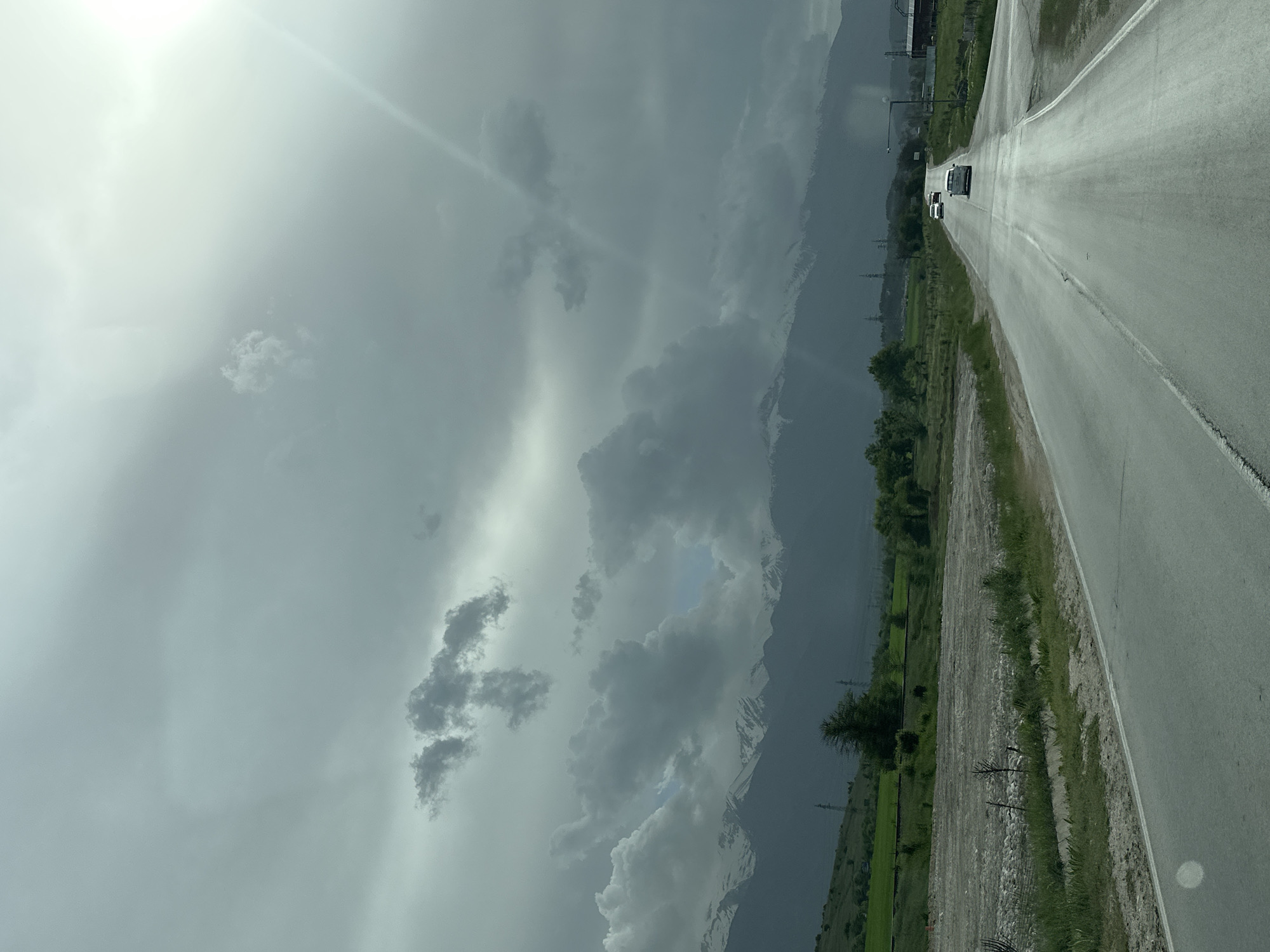 The image shows a road with cars in the distance, alongside a green field, with mountains and a cloudy sky. The location is Unknown, Unknown.