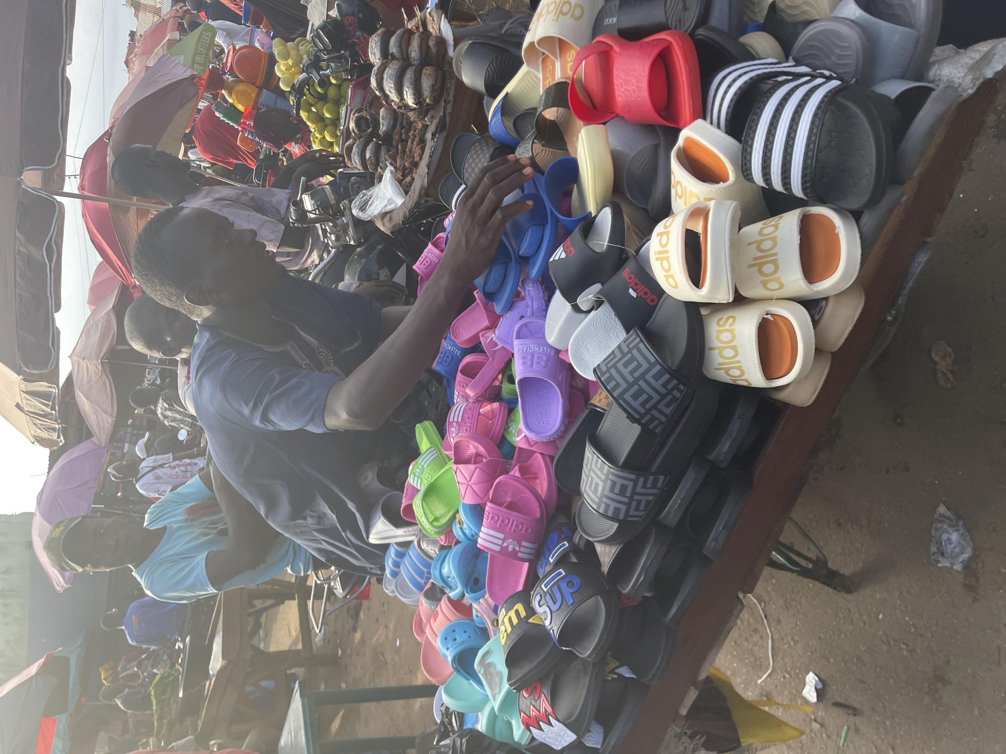The image captures a vibrant market scene in Akwanga, Nigeria. A man wearing a blue shirt is sorting through a large pile of colorful sandals displayed on a wooden table. The sandals are various styles and brands, including Adidas, Balenciaga, and others. The market is busy, with people and various goods under umbrellas visible in the background. Other items for sale, like fruits and possibly coconuts, are also seen in the background, indicative of a bustling marketplace environment. The ground appears to be dirt or paved and litter can be found. The overall impression is that of a lively, open-air market characteristic of many African cities.