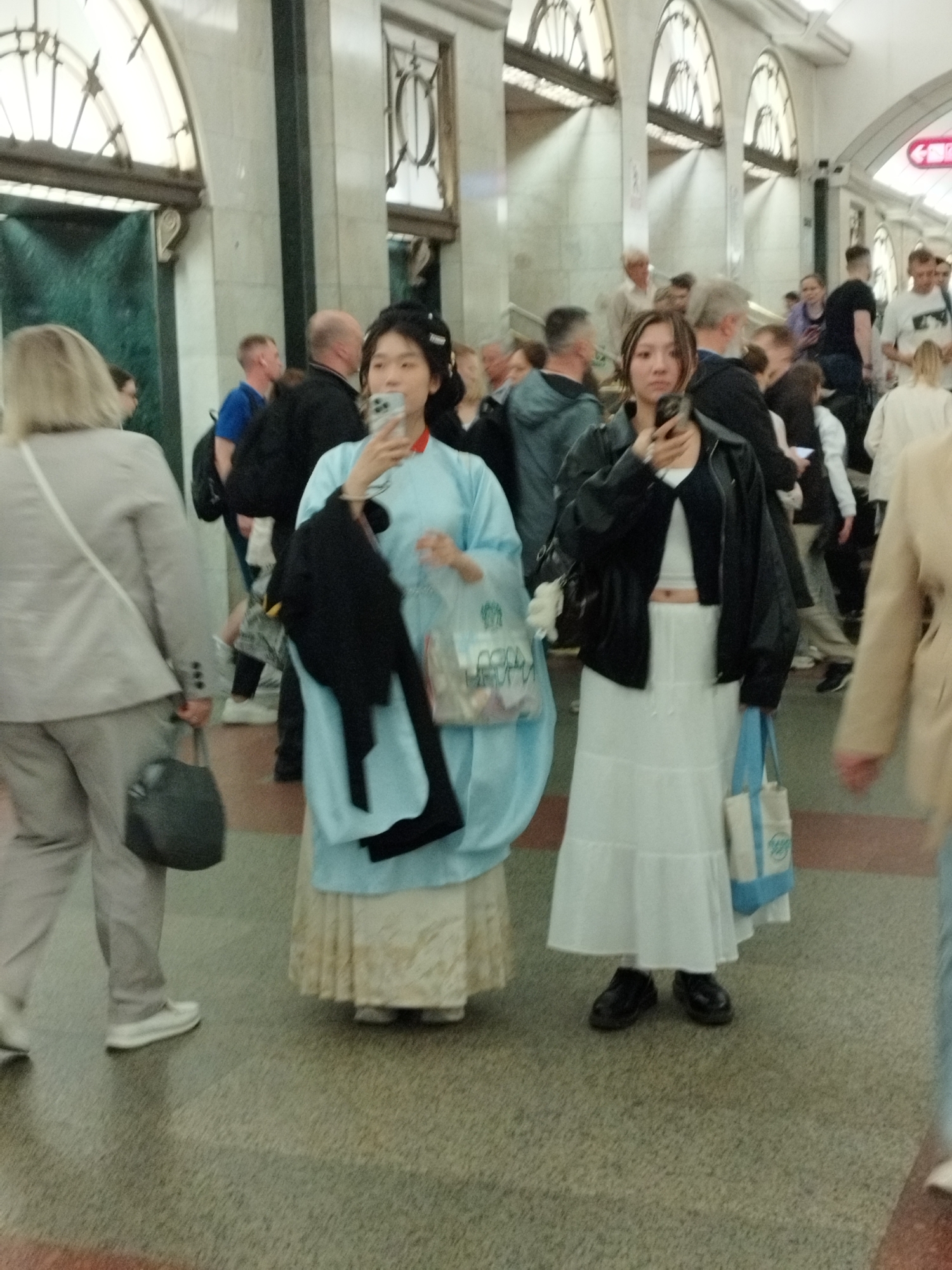 The image shows a crowded indoor environment, likely a subway station in Saint Petersburg, Russia, given the architectural style with arched ceilings and marble-like walls. Two young women are prominently featured in the foreground, each holding a smartphone.
The woman on the left is wearing a light blue traditional-style garment over a cream-colored layered skirt. She is holding a black jacket in her left arm and a transparent plastic bag in her right hand. She is looking at her phone.
The woman on the right is wearing a black leather jacket over a white crop top and a long white tiered skirt. She is carrying a blue and white tote bag over her left shoulder and is also looking at her phone. She is wearing black shoes.
Several other people are visible in the background, blurred due to the focus on the foreground subjects. They appear to be commuters, with some walking and others standing. The floor is a speckled pattern, and there are green-tiled doors or entrances in the background. The lighting suggests an indoor public space, possibly illuminated by artificial lights.