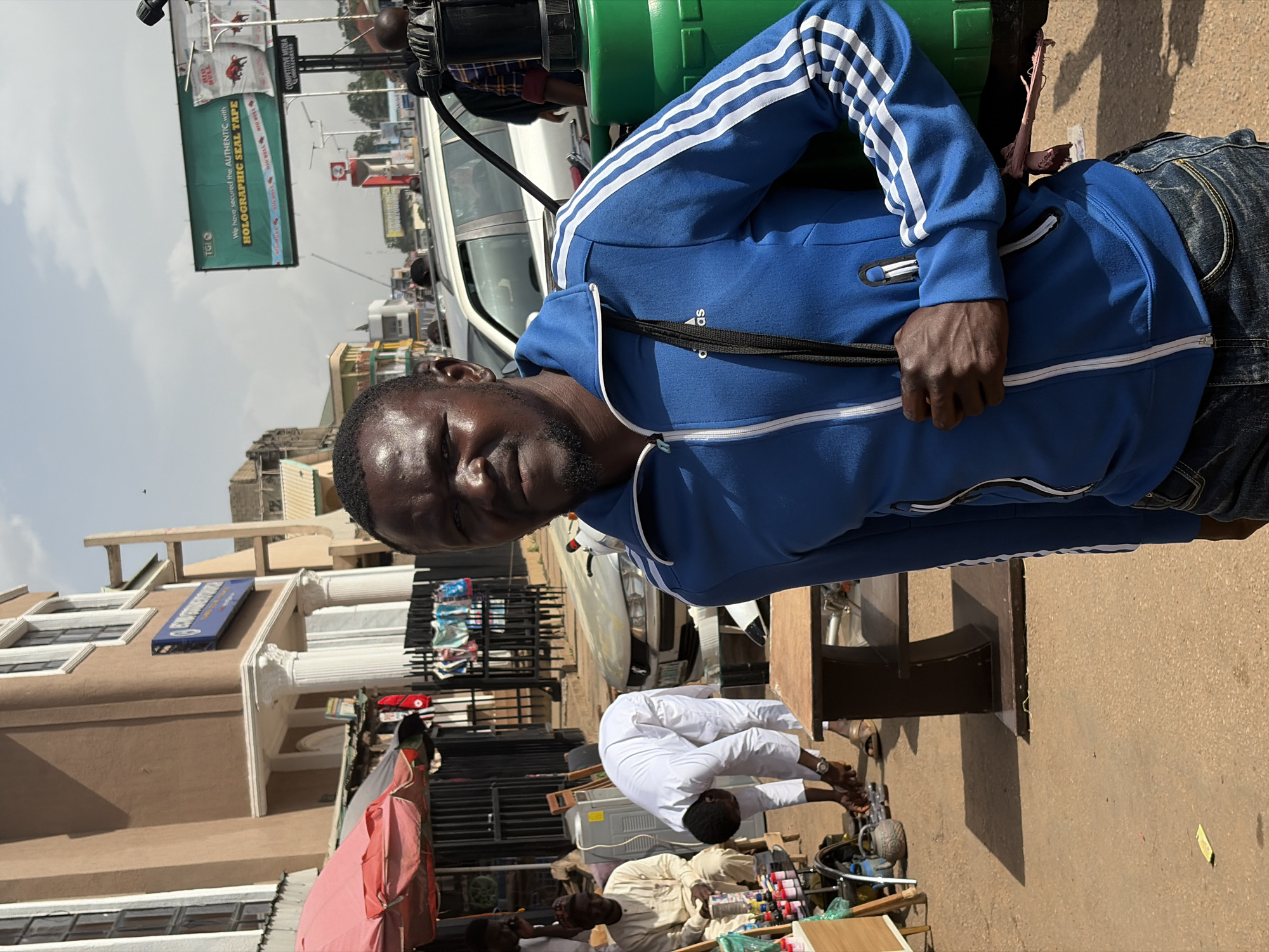 A man in a blue Adidas tracksuit and jeans stands in the foreground of a street scene in Jos, Nigeria. He is looking towards the camera with a neutral expression. Behind him, several other individuals are visible. One person in a white robe is bent over, seemingly examining something on the ground near a table. Another person in a cream-colored robe is seated. In the background, there are buildings, vehicles, and a large billboard. The environment appears to be a busy urban street, possibly a marketplace. The lighting suggests it is daytime, with a clear sky overhead.