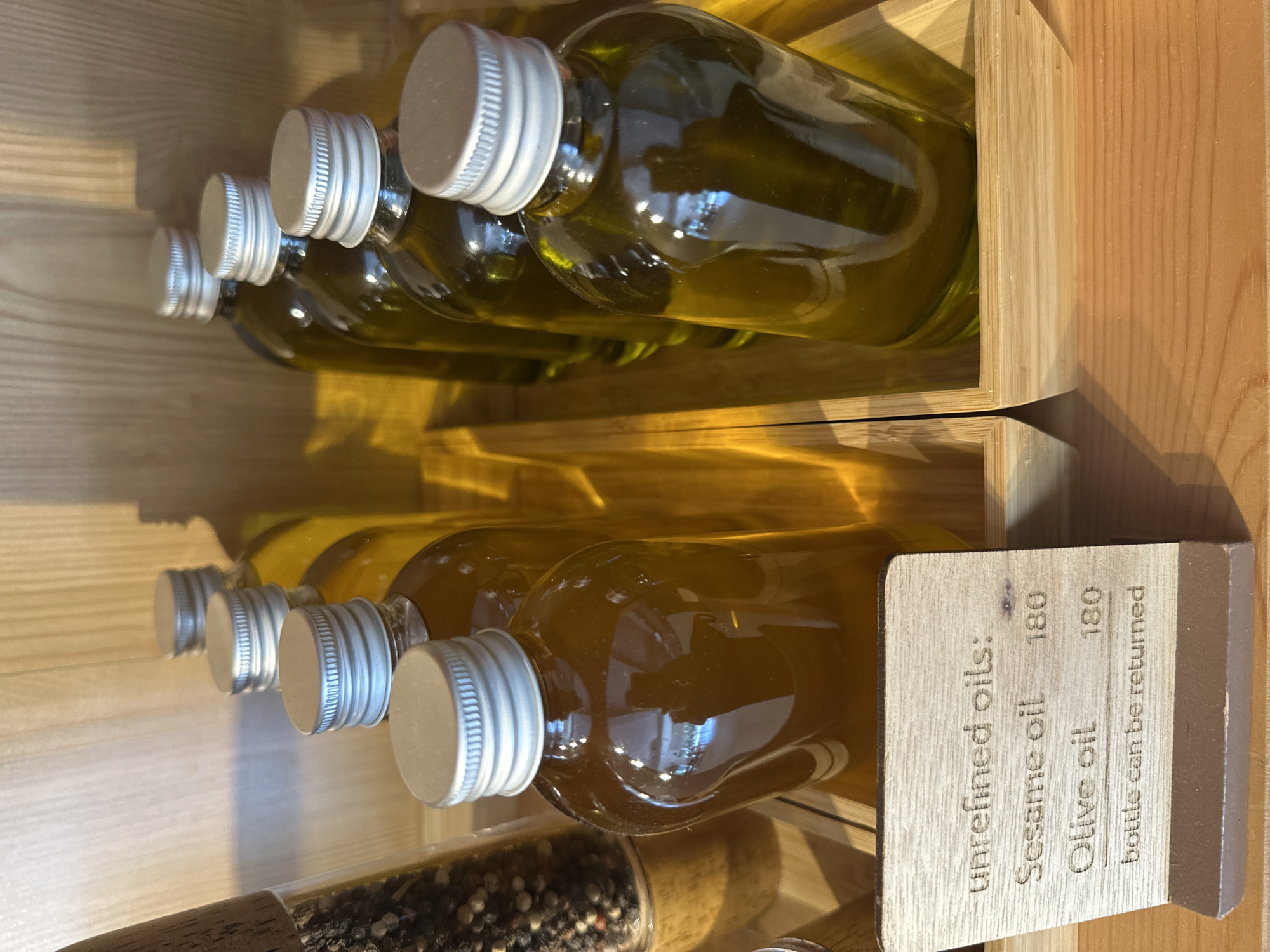 In the city of Koh Phangan, Thailand, within a retail environment, several glass bottles filled with amber-colored liquid are displayed in wooden crates. The bottles have silver screw-on caps. A small, wooden sign is positioned in front of the bottles, displaying text that reads "unrefined oils:", "Sesame oil 180", "Olive oil 180", and "bottle can be returned". The background is a wooden surface. To the left, a glass container with peppercorns is partially visible. The lighting suggests an indoor setting.