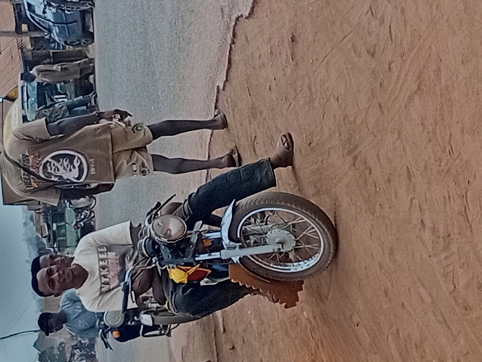 The image depicts a roadside scene in Ayangba, Nigeria. In the foreground, a light-skinned male, approximately 20-30 years old, is seated on a motorcycle, facing slightly left of center and smiling towards the camera. He wears a light-colored t-shirt with dark text, possibly reading "WAKEEL USMAN," and dark trousers. His left hand rests on the motorcycle's handlebar. The motorcycle is light blue/silver with a chrome headlight and spoked wheels, parked on a reddish-brown dirt shoulder.
To the right and slightly behind the seated male, a dark-skinned male, also approximately 20-40 years old, walks away from the camera, facing towards the right. He wears a tan t-shirt with a prominent graphic on the back featuring a stylized face and the text "HELP STAR." He also wears tan shorts and open-toed sandals. He holds a small, light-colored object in his left hand.
The background shows a paved road with multiple vehicles, including other motorcycles, cars, and a light-colored van or truck. Several other individuals are visible, some standing by the road, others seemingly in transit. Structures with reddish-brown roofs are present in the distance. The sky appears overcast. The immediate ground in the foreground is unpaved, consisting of reddish-brown dirt, indicating a roadside area adjacent to a paved street.