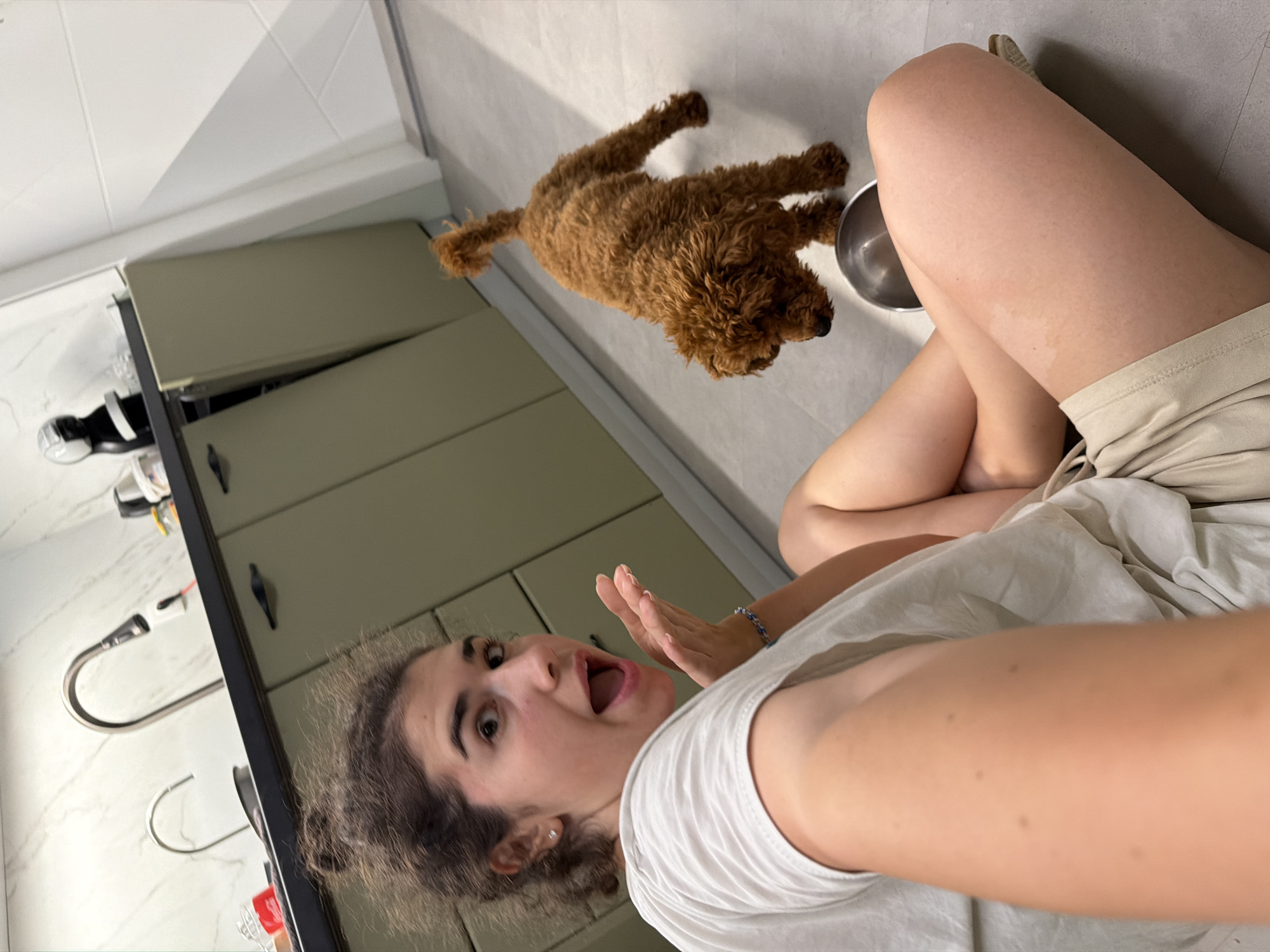 A woman with curly brown hair tied up in a bun is sitting on the floor of a modern kitchen, looking at the camera with her mouth open in surprise. She is wearing a white t-shirt and beige shorts. To her right, a small, fluffy brown poodle is standing with its tail up, looking down at a silver bowl on the floor. The kitchen has light gray tiled floors and sage green cabinets. In the background, a chrome faucet is visible above a sink against a marble-tiled backsplash. The image appears to have been taken in Sofia, Bulgaria, based on the context provided.