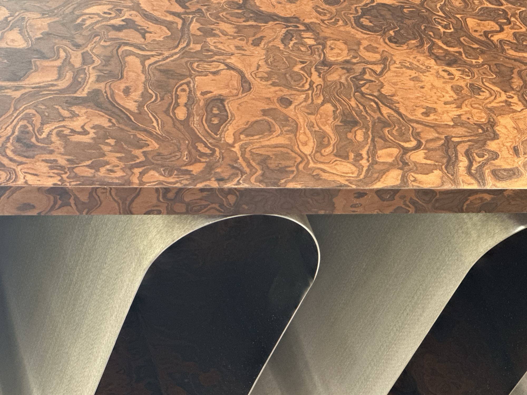 The image depicts the corner of a table with a burl wood top and metal legs. The table legs are curved and appear to be made of brushed metal. The background is blurred, but the setting is indoors. The location is Milan, Italy.