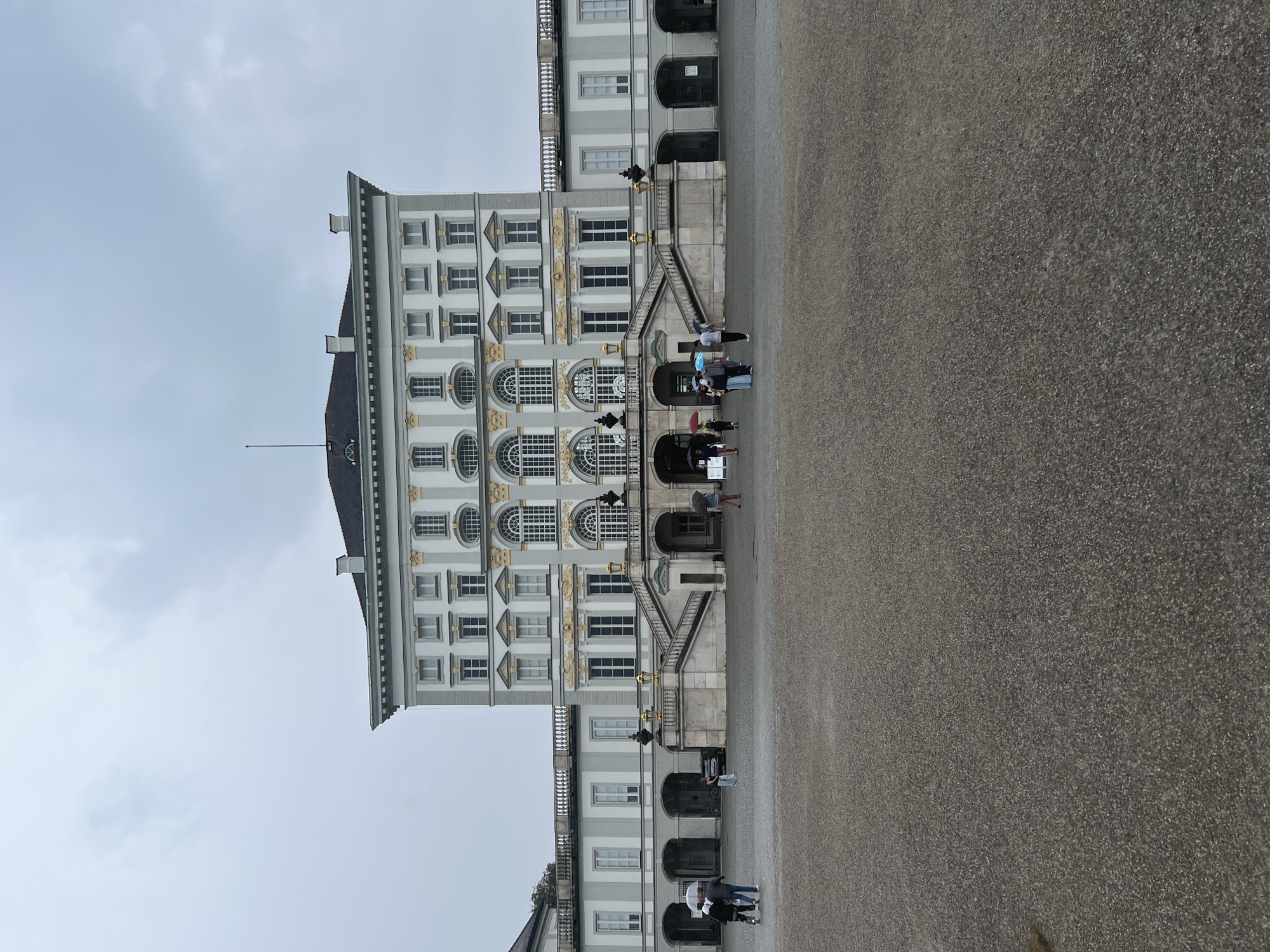 The image shows a wide, gray, gravel-covered courtyard in front of a grand, pale gray, multi-story building with a dark roof. The building features a symmetrical facade with numerous windows, some arched and others rectangular, and decorative gold accents. A prominent set of wide, stone stairs with ornate balustrades leads up to the central entrance. Several people are visible in the courtyard and on the steps, some holding umbrellas, suggesting it is a cloudy or rainy day. The architecture suggests a palace or a significant historical building, consistent with Nymphenburg Palace in Munich, Germany, which is known for its expansive courtyards and Baroque style. The sky is overcast and a pale gray, with no discernible clouds.