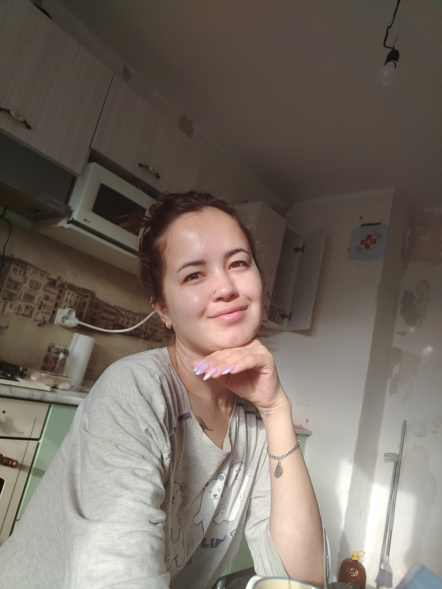 The image is a selfie of a young woman with dark hair pulled back in a bun. She is wearing a grey t-shirt with a pattern of bears. Her right hand is propped under her chin, and she has long, light purple fingernails. She is wearing a silver bracelet on her left wrist. The woman is smiling slightly and looking at the camera. The background appears to be a kitchen. There are cabinets on the wall, one of which is open. A microwave is visible above a counter. A patterned backsplash is behind the counter. Sunlight is streaming into the room from the right, casting shadows. The overall impression is of a casual, indoor scene. The location is identified as Noviy, Russia.