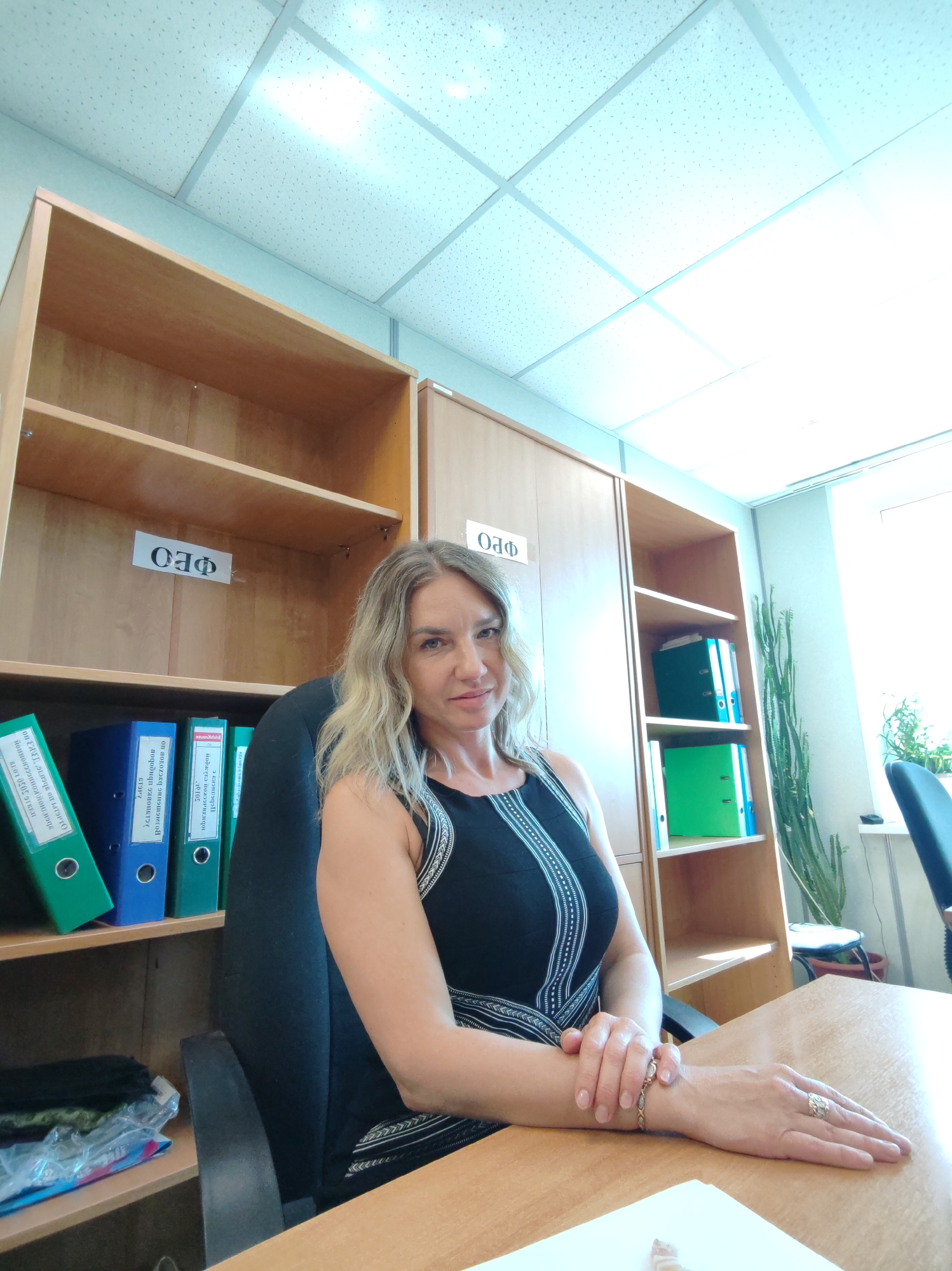 A medium shot shows a blonde woman sitting at a desk in an office. The woman is wearing a black sleeveless dress with white detailing on the front. She has her arms crossed on the desk. On her right wrist is a gold bracelet, and on the ring finger of her right hand is a silver ring with a large stone. Behind her is a wooden bookcase with multiple shelves. The shelves contain several binders and some personal items. On the top shelf of the bookcase is a sign that reads "ОФО" in Cyrillic script. Behind the bookcase is a window with a potted plant. The office has a drop ceiling with fluorescent lights.