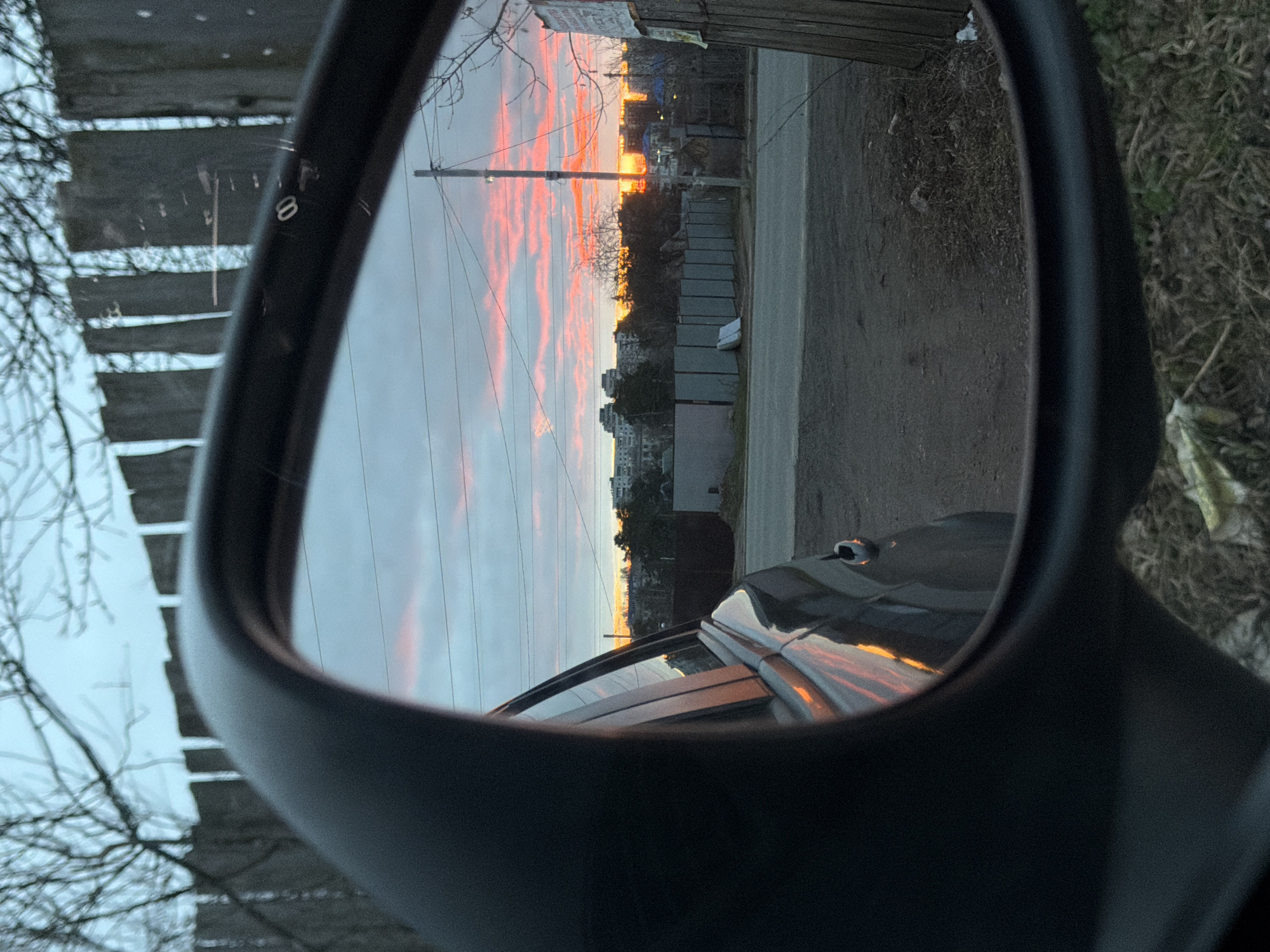 The image is a unique perspective shot, captured through a car's side-view mirror, revealing a vibrant sunset or sunrise over a semi-urban landscape in Chaikovsky, Russia. The sky, as reflected in the mirror, is painted with dramatic streaks of bright orange and pink clouds, indicating either dawn or dusk, with the low sun casting a warm, golden glow across the scene.
Reflected in the mirror is a landscape featuring power lines draped across utility poles, several multi-story buildings in the distance, and a road or path extending into the background. Bare tree branches are visible in the foreground, suggesting a late autumn or early spring season. A rustic wooden fence or structure is partially visible on the left side of the reflected scene. Parts of the car's own body are also reflected, showing the warm hues of the sky.
The black frame of the car's side mirror occupies a significant portion of the image, framing the reflection. A small, white, circular "8" is faintly visible on the top-left section of the mirror's housing. No people are visible in the image. On one of the utility poles in the reflection, a blurry piece of paper is attached, but its text is illegible. The scene conveys a quiet observation of a natural spectacle from inside a vehicle.