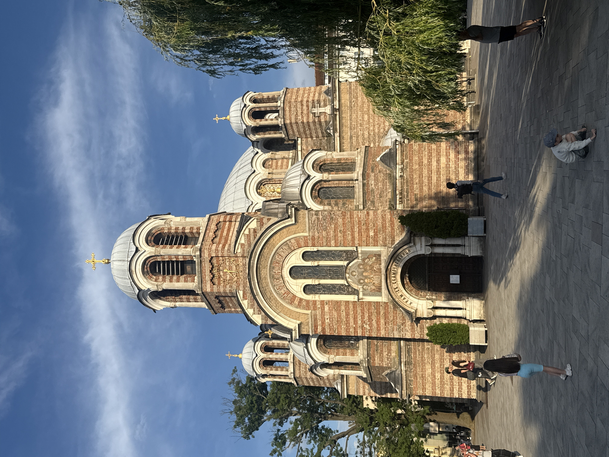 The image depicts the majestic Church of the Holy Seven Saints (Sveta Sedmochislenitsi) in Sofia, Bulgaria, on a clear, sunny day. The large Eastern Orthodox church dominates the left side of the frame, constructed from alternating bands of reddish-brown and lighter cream-colored bricks, creating a distinctive striped pattern. It features several grey domes, each adorned with a golden cross at its apex. Ornate arched windows with dark grilles and decorative stone carvings are visible across the facade, with frescoes or mosaics above some arches. Prominently displayed above the main entrance is Cyrillic text that reads "ХРАМ СВ. СЕДМОЧИСЛЕНИЦИ" (Church of the Holy Seven Saints).
In front of the church, a paved plaza extends to the right, bathed in sunlight. Scattered shadows from trees and the church suggest a late morning or early afternoon setting. Several people are present in the plaza:
- One individual in blue shorts and a white top is seen stretching or lying flat on the ground with arms and legs extended.
- Nearby, a person, possibly a child, wearing a light hoodie and a blue cap, is crouched down.
- Further back, a person in dark shorts and a grey shirt stands beneath a lush green tree with weeping branches.
- Another person with dark hair, wearing a light shirt and jeans, walks away from the camera towards the church entrance.
- Two other figures are visible closer to the church entrance, one carrying a red bag.
The sky is a vibrant blue with sparse, wispy white clouds, and verdant trees frame the top and right edges of the scene. The overall impression is one of a serene public space with casual activity around a beautiful historical and religious landmark.
