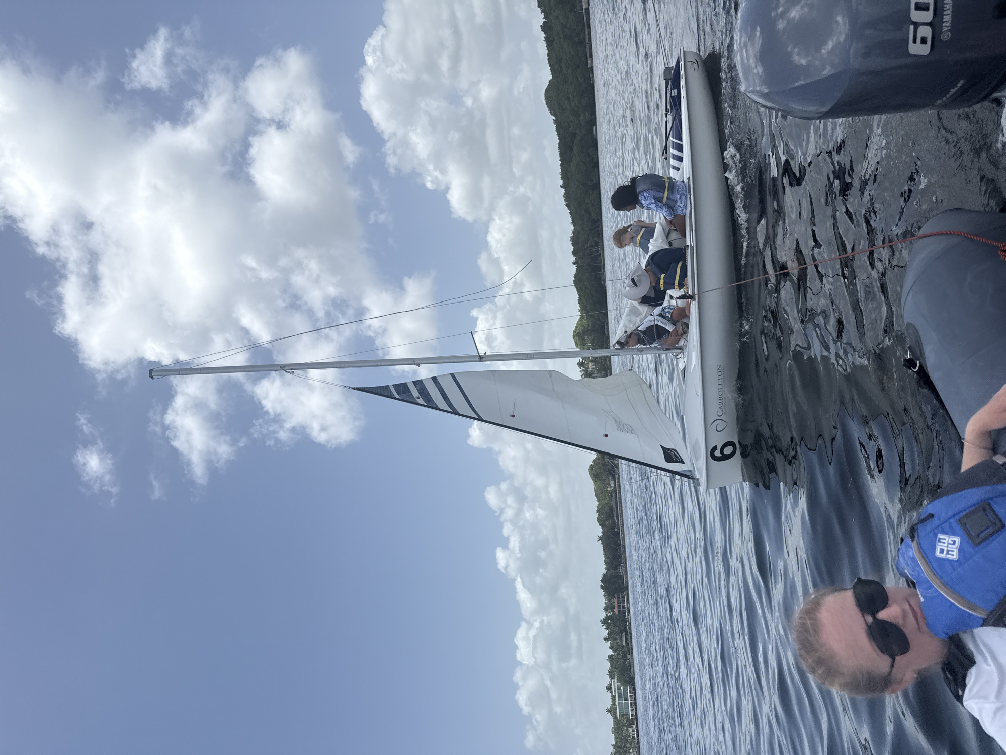 An aerial medium shot captures a bright, sunny day on the water in Miami, Florida. The scene is dominated by a sailboat numbered '6' with "CARROLLTON" written on its hull, carrying four people. In the foreground, closer to the viewer, another person wearing sunglasses and a life vest is visible, seemingly in another boat. The water is a deep blue, with gentle ripples reflecting the sky and clouds. A lush green shoreline with palm trees and houses lines the horizon, suggesting a tropical or subtropical environment. The sky is a clear blue, punctuated by fluffy white clouds. The overall atmosphere is one of leisure and outdoor activity on a beautiful day.