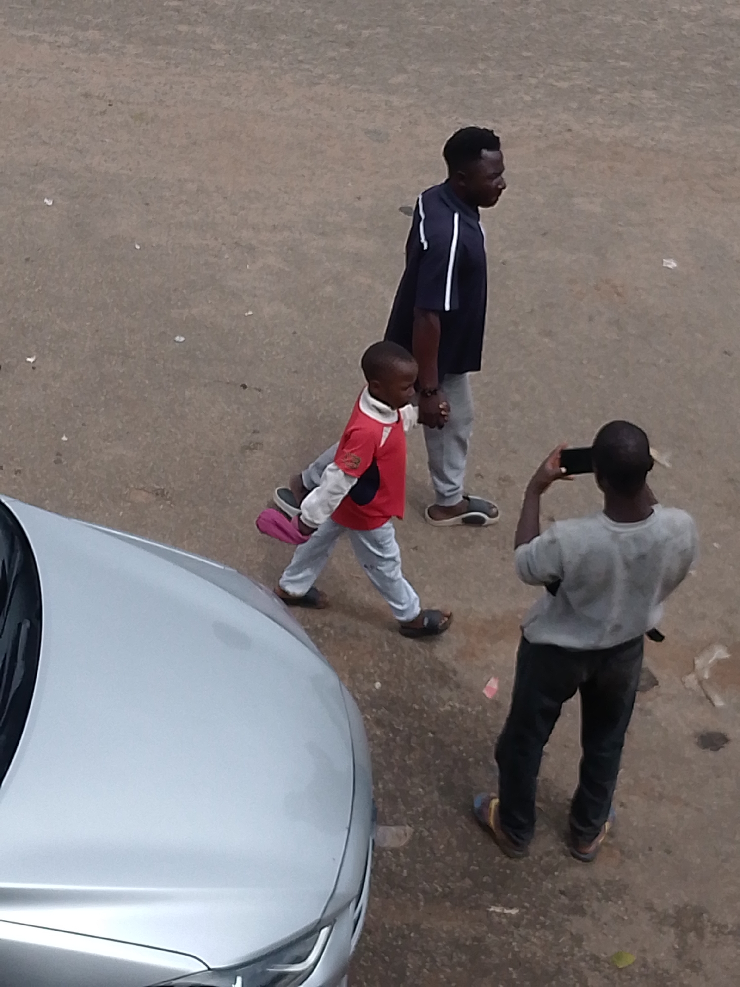 In Bukuru, Nigeria, on a paved surface, an adult male in a dark blue shirt with white stripes walks hand-in-hand with a young boy wearing a red shirt and grey pants. The boy is carrying a pink object. Behind them, another adult male stands with a phone, appearing to take a photo. A silver car is partially visible on the left side of the frame. The ground is concrete with scattered debris.