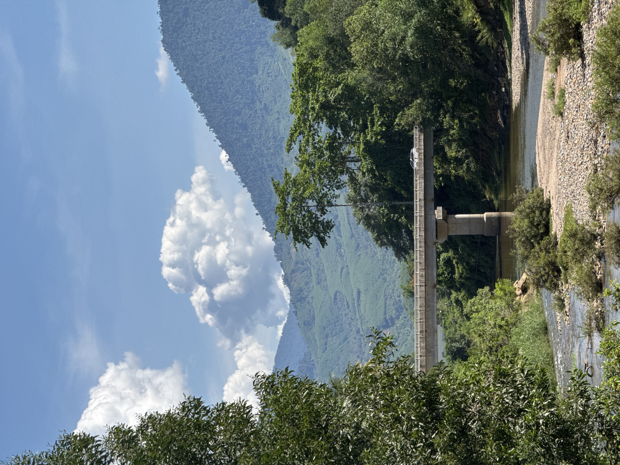 An outdoor, eye-level shot captures a bridge spanning a river in Nam Khanh Vinh, Vietnam. The bridge is made of concrete and supported by substantial pillars, with a car visible driving across it. Lush green trees flank the river, with a dense forest and rolling, verdant mountains forming the background. The sky above is a clear blue, dotted with fluffy white clouds. The river flows over a rocky bed, with some vegetation growing along its banks. The overall scene suggests a tranquil, natural landscape enhanced by human infrastructure.