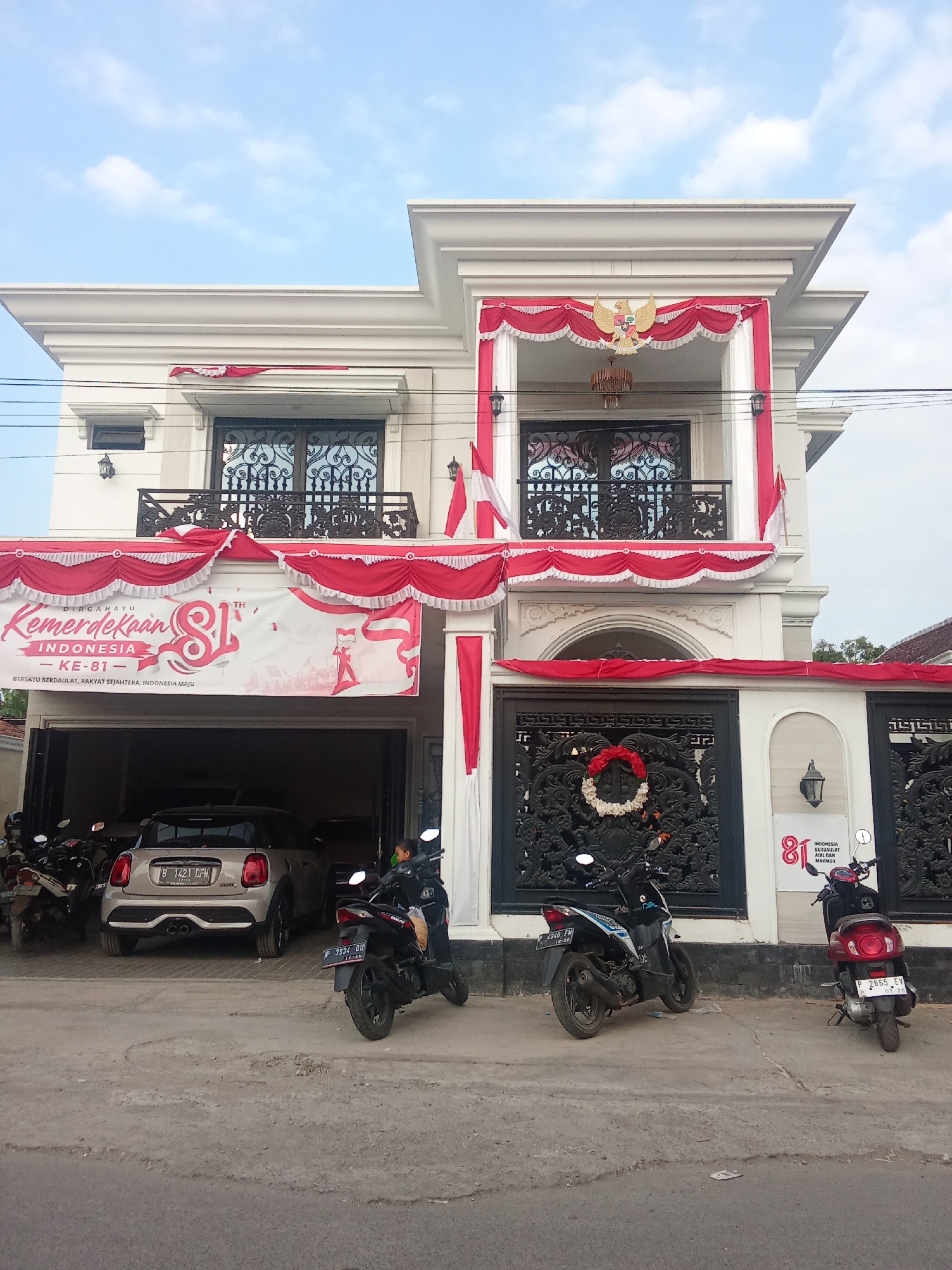 A two-story building decorated with red and white banners and flags, celebrating Indonesia's 81st Independence Day. The building has a modern architectural style with white walls and intricate dark metal railings on the balconies. A large banner with "Dirgahayu Kemerdekaan Indonesia Ke-81" is prominently displayed. The ground floor features a garage with a silver Mini Cooper and several motorcycles parked outside. The street in front of the building is paved and shows signs of wear. The sky is partly cloudy.