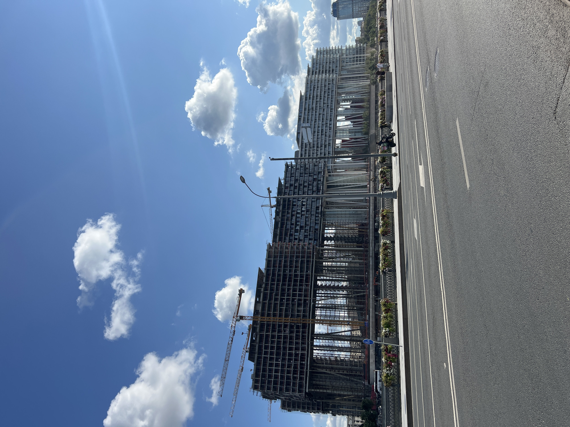 An outdoor, daytime, eye-level shot captures a large building under construction in Moscow, Russia, with a clear blue sky dotted with fluffy white clouds. The building, which appears to be a multi-story complex, is partially enclosed in scaffolding and features numerous support columns. Two cranes are visible on top of the structure, indicating ongoing construction. In the foreground, a paved road with white lane markings runs horizontally across the frame. A sidewalk with decorative planters and a metal fence runs parallel to the road. A few people are visible on the sidewalk, along with a scooter. The environment suggests an urban setting with modern architecture.