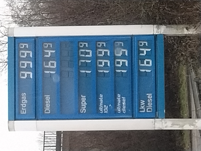 The image displays an electronic gas station price board, rotated 90 degrees counter-clockwise, against a blurred background of bare trees or bushes, suggesting an outdoor setting during daylight hours with dull or overcast weather. No people are visible.
The blue price board features white digital numbers and white text labels for various fuel types, consistent with a gas station in Germany (Saarlouis context provided).
The visible text and corresponding prices are:
* **Erdgas** (Natural Gas): 9.999
* **Diesel**: 1.649
* **Super** (Standard gasoline, typically Super 95): 1.709
* **ultimate 102** (Premium gasoline, 102 octane): 1.999
* **ultimate diesel** (Premium diesel): 1.959
* **Lkw Diesel** (Truck Diesel): 1.649
The prices are likely displayed in Euros per liter, a standard format in Germany, indicated by the three decimal places with the last digit being smaller or implied as fractional. The board is bordered by a white frame.