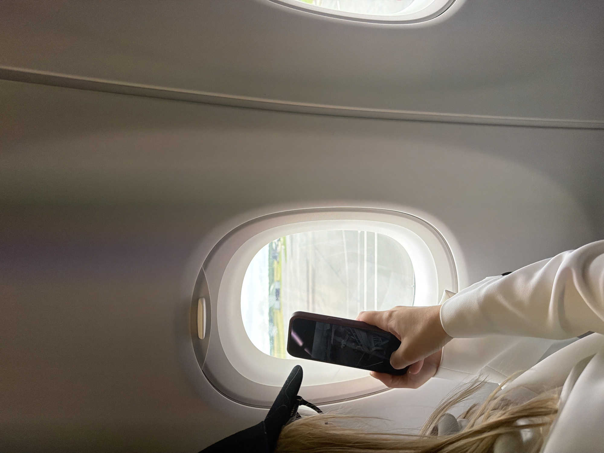 This image provides an intimate view from an airplane window seat, likely during daytime. The primary subject is a person, seen from the chest down, holding a dark-cased smartphone up to the oval-shaped aircraft window. Their right hand grips the phone, which appears to be dark or powered off, reflecting the bright exterior light. Long, light-colored hair of the person cascades on the right side of the frame, and a leg or foot wearing a black shoe is visible near the bottom left of the window.
The interior of the airplane cabin is characterized by smooth, light grey walls and ceiling, featuring a subtle horizontal groove or light strip running across the upper section. Through the window, a bright outdoor scene is visible, showing elements of green foliage and light-colored structures, suggesting an airport environment or landscape. The illumination from outside indicates it is daytime. The activity captured is a common one during travel: a passenger interacting with their phone while looking out the window, possibly taking a photo or video. Given the location context, this scene likely takes place at or near Tillé, France. No discernible text is visible in the image.