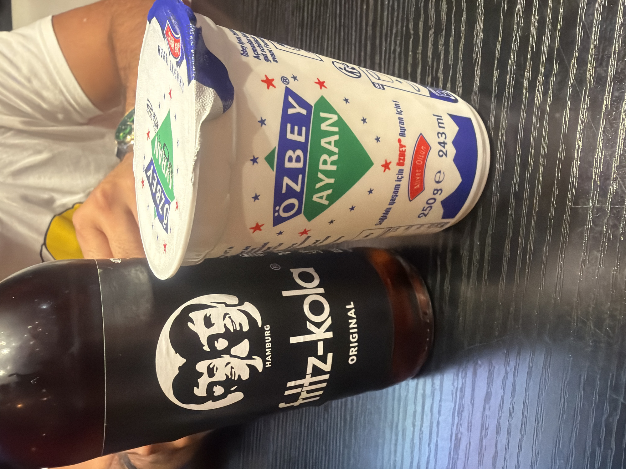 Two beverages, a bottle of Fritz-Kola and a cup of Özbey Ayran, are displayed on a dark, textured surface, likely a table. The Fritz-Kola bottle is black with white text and graphics, featuring the brand name "fritz-kola" and the word "ORIGINAL". It also shows the "HAMBURG" location and a stylized image of two men's faces. The Özbey Ayran cup is white with blue and green branding, displaying "ÖZBEY AYRAN" prominently. Text in Turkish and German is visible on the cup, indicating "Sağlıklı yaşam için ÖZBEY Ayran için!" (For a healthy life, for ÖZBEY Ayran!) and "Açmadan be... gut schü... avant l'em...". The cup also indicates its volume as "250 g e 243 ml". The background shows a person wearing a white t-shirt with yellow accents on their left shoulder, with their arms visible, suggesting they are seated at the table. The lighting is somewhat dim, highlighting the products. There is no specific indication of the city of Duisburg, Germany, in the image itself.