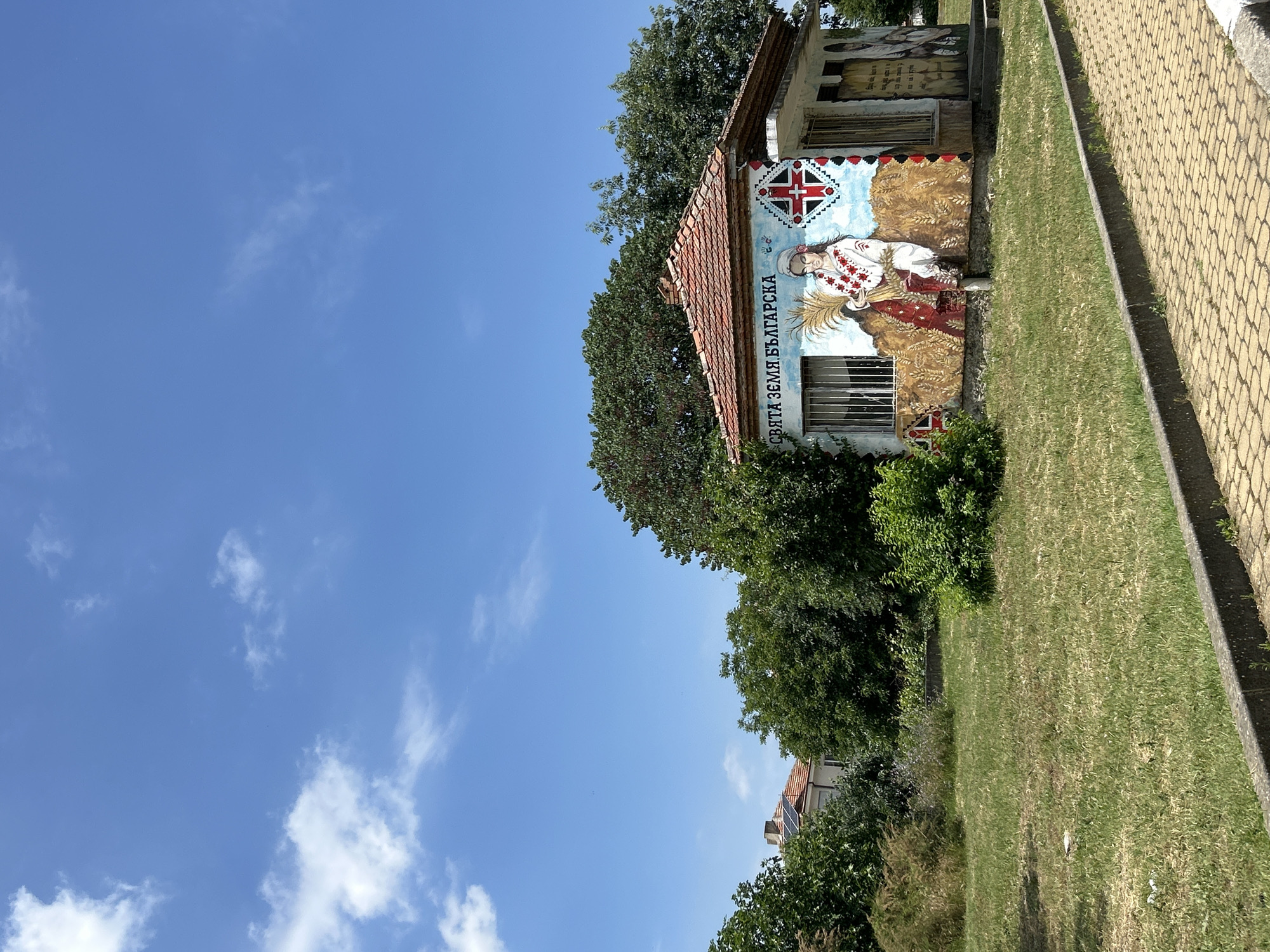 A single-story building in Aheloy, Bulgaria, is adorned with vibrant murals. The building has a terracotta tiled roof and is surrounded by green grass and trees. A paved walkway runs in front of the building. The mural on the front wall depicts a woman in traditional Bulgarian clothing holding a sheaf of wheat, with a stylized Bulgarian flag on the right side of the wall. Text above the woman reads "SVYATA ZEMYA BALGARSKA" (Holy Bulgarian Land). Another mural on the right side of the building shows a man with a shield and spear. The sky above is blue with scattered white clouds. The scene appears to be a sunny day.
