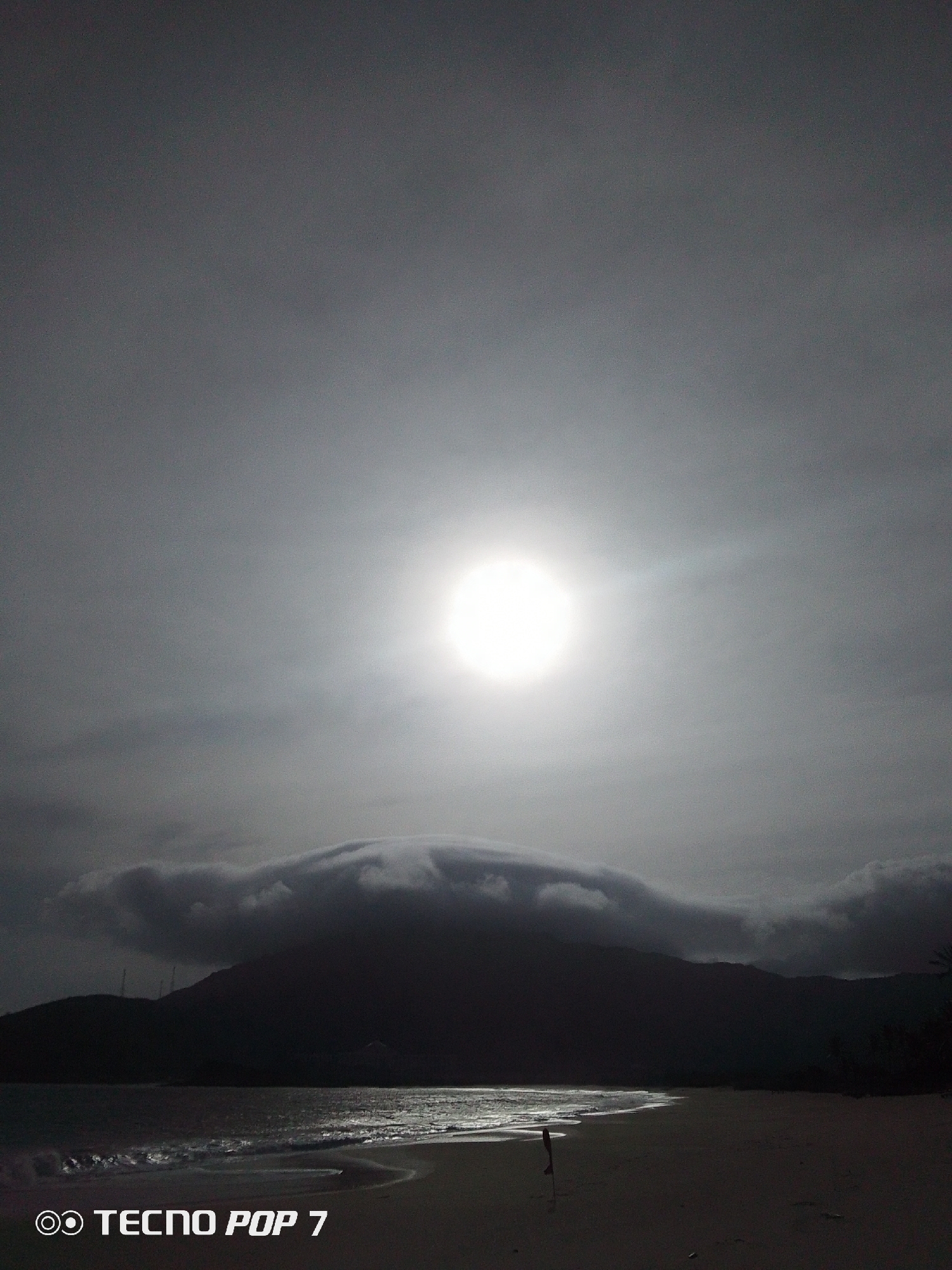 The image shows a monochrome view of Playa Caribe in Pedro González, Venezuela, under a bright sun obscured by clouds. The beach extends in the foreground, where waves are visible. In the distance, a dark mountain range is partially covered by low-lying clouds. A single figure stands on the beach. The photo was taken with a TECNO POP 7 phone, as indicated in the bottom left corner of the image.