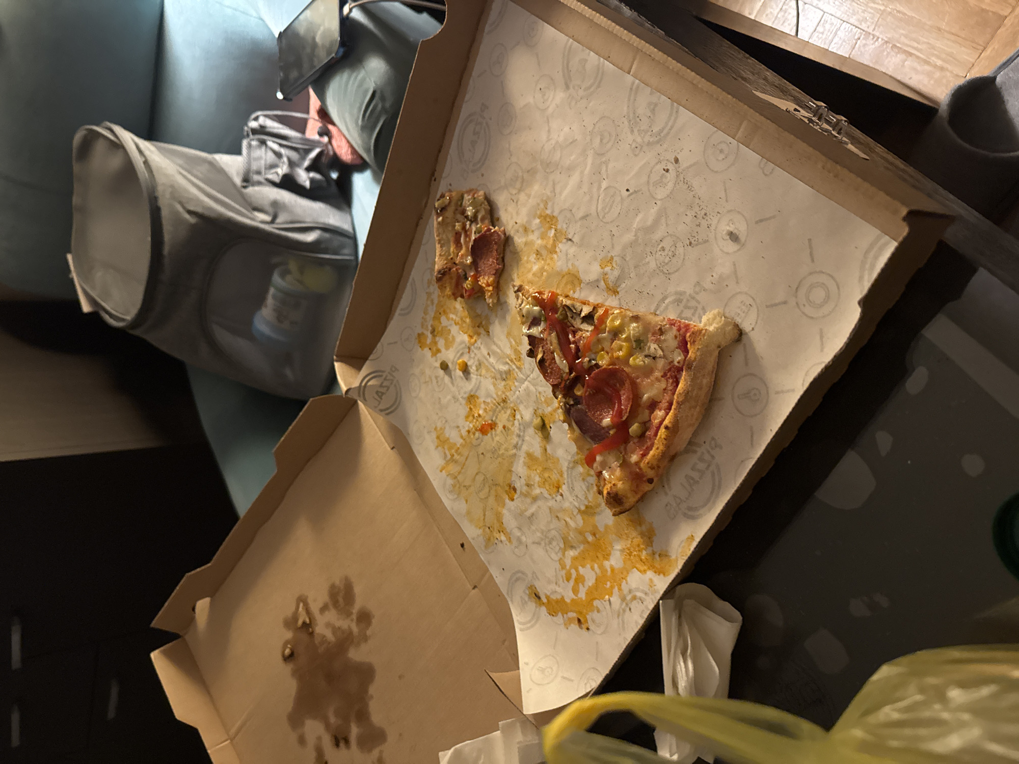 The scene depicts an open pizza box containing one and a half slices of pizza. The pizza has pepperoni, mushrooms, corn, and red peppers as toppings. The box is stained with grease and sauce. Adjacent to the box is a grey insulated bag and a partly visible cup. The image suggests a casual, indoor setting. It's impossible to determine the exact location in Sofia, Bulgaria, or any people, actions, or interactions involved, without additional information.
