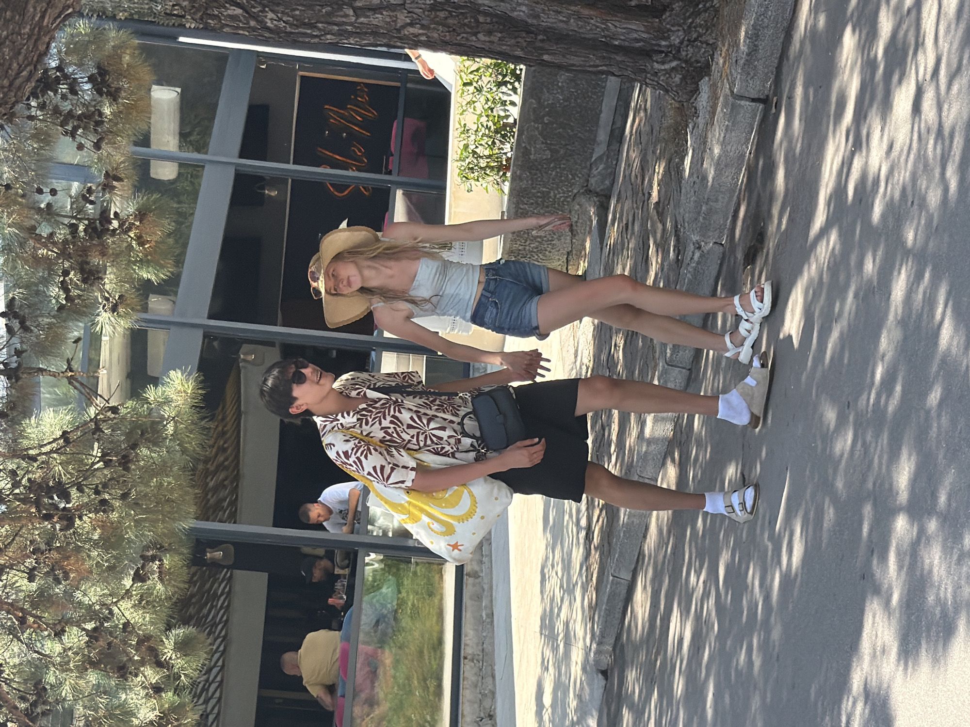 The image shows two people walking on a sidewalk next to a building with large windows. The person in the foreground is a man wearing a patterned short-sleeved shirt, black shorts, sunglasses, and carrying a tote bag with a yellow octopus design. He is looking up and to his right, with a slight smile. The person behind him is a woman wearing a straw hat, a light blue tank top, and denim shorts. She is walking with her arms outstretched and has a peace sign gesture with her left hand. Both individuals are wearing sandals.
The scene appears to be outdoors during the day, with sunlight casting shadows on the ground. A tree with sparse foliage is in the foreground, obscuring part of the view. Through the building's windows, glimpses of what appears to be a restaurant or cafe interior can be seen, with patrons and tables. The building's facade has a sign with orange lettering, though the text is partially obscured and difficult to read clearly, but it appears to say "Belle Ami". The overall atmosphere suggests a casual, warm-weather day, possibly in a tourist area. The location is identified as Budva, Montenegro.