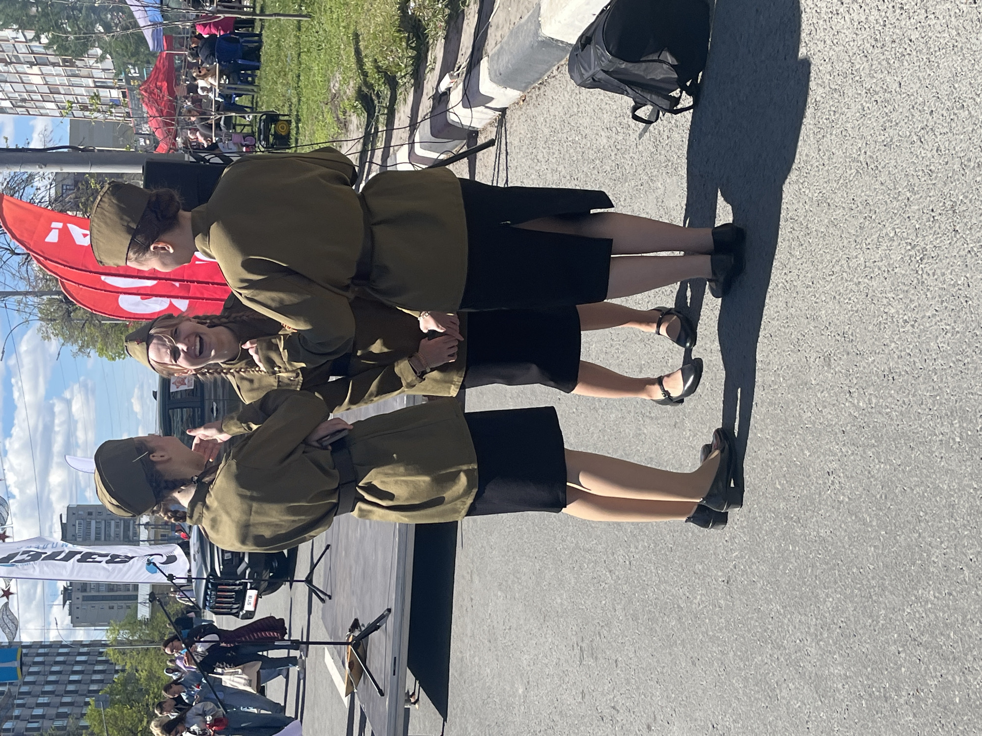 Three women are standing on a stage in Ulyanovsk, Russia, likely at a public event. They are dressed in matching khaki military-style jackets and hats, with black skirts and shoes. The women appear to be performers, as they are standing near microphones and musical instruments on the stage. Behind them are red banners and a small crowd of people watching the event. The background shows city buildings under a partly cloudy sky. The foreground reveals a grey pavement surface.