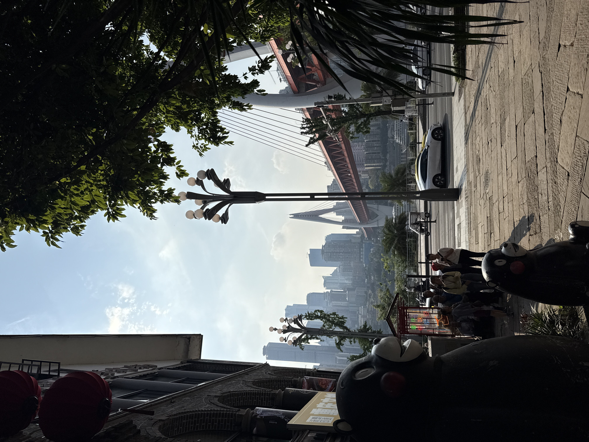 A ground-level, rotated view shows a street scene. In the foreground, two dark, cartoonish figures stand to the right. Behind them, people are seated at tables. The city skyline rises in the background, featuring a large bridge. Overhead, a streetlight with multiple bulbs extends to the left of the frame, positioned against a light blue sky with some clouds. A glimpse of architecture is seen along the lower left, including a sign. Cars and other signs of urban life can be seen on the street level. The location appears to be in the Nan'an District, China.