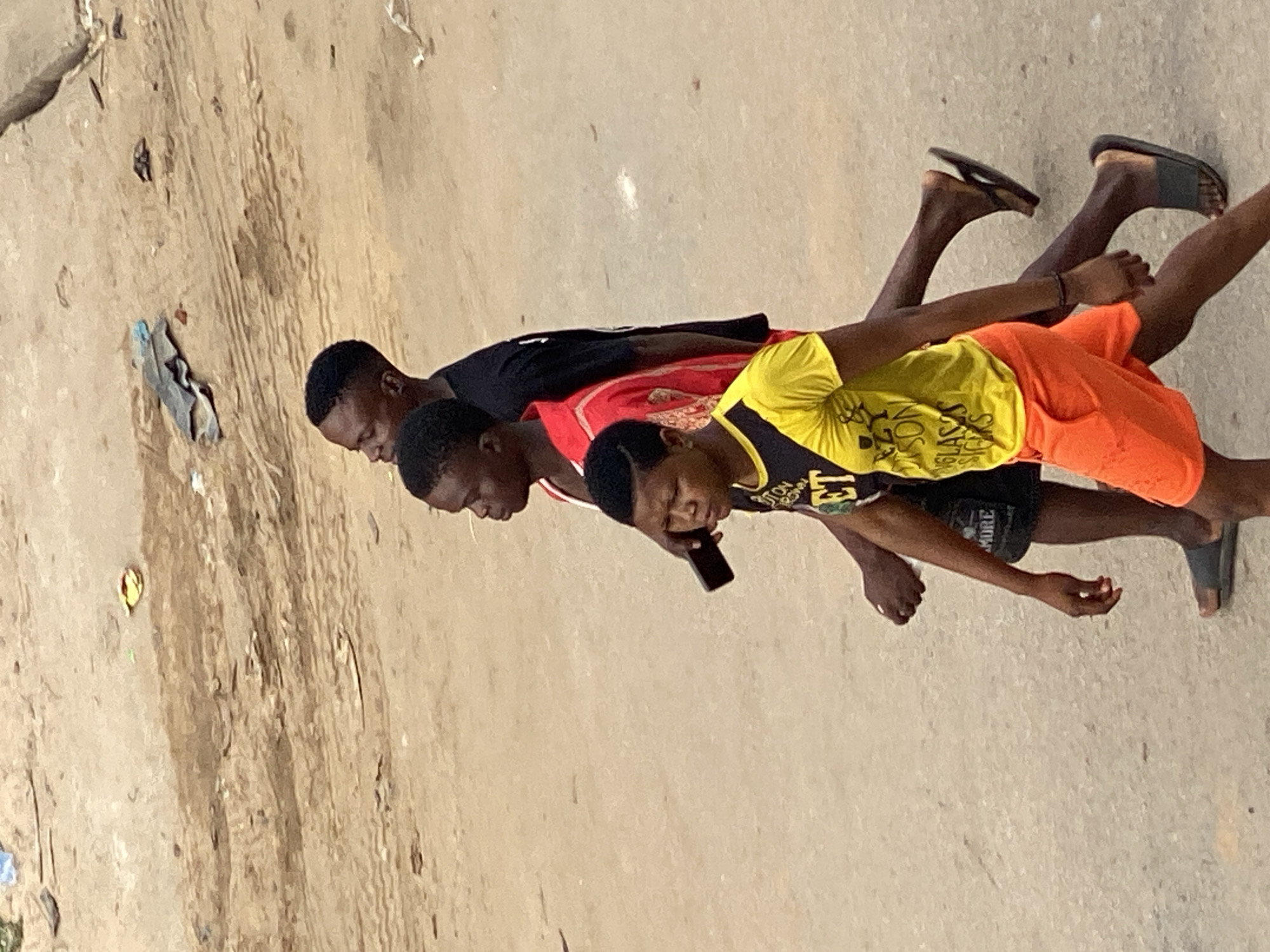 Three young men are walking on a dusty street in Fegge, Nigeria. The man closest to the viewer is wearing a yellow shirt and orange shorts and holding a black mobile phone. The second man is wearing a red shirt, and the third man is wearing a black shirt. All three are wearing flip-flops. The road is sandy and there is litter on the side.