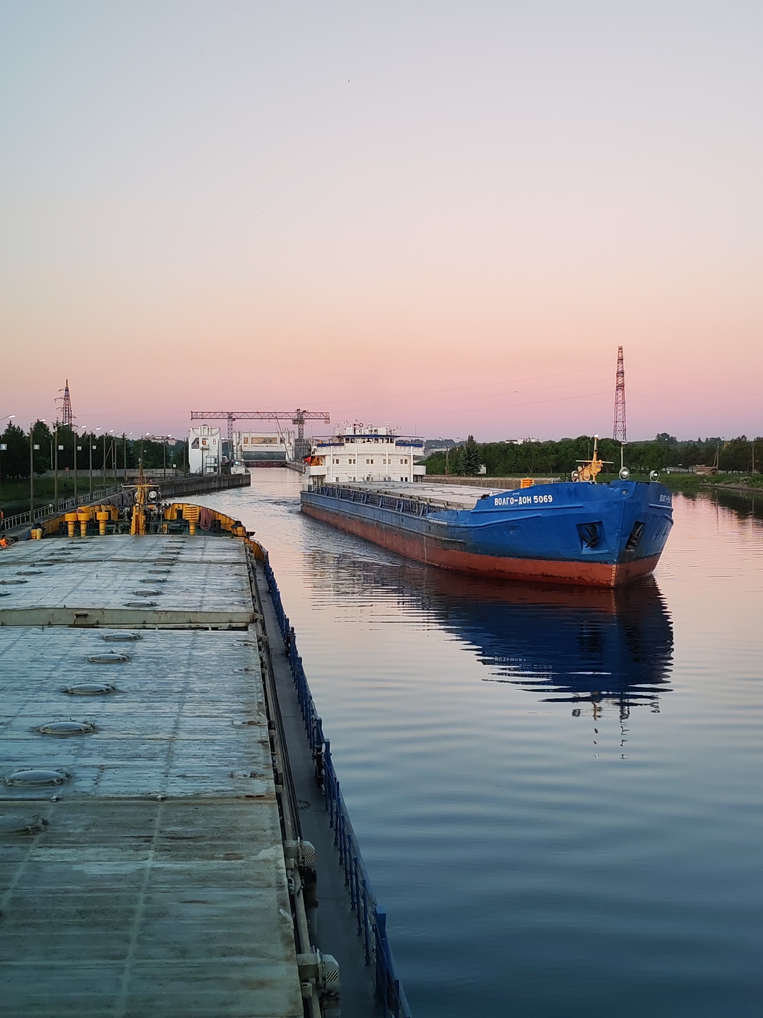 The image captures a tranquil industrial waterway, likely a canal or river, at dawn or dusk in Unknown, Unknown. In the foreground, the deck of a large, flat-topped cargo vessel, possibly a barge, occupies the left side, featuring numerous circular metal hatch covers and yellow-painted mechanisms. A blue railing separates the vessel from the water. In the mid-ground, a blue and red cargo ship, bearing the name "ВОЛГО-ДОН 5069" (Volgo-Don 5069) on its bow, navigates the calm water, heading towards the viewer. Its reflection is distinct in the water. The background reveals a series of concrete structures, indicative of a canal lock system, flanked by industrial buildings and a large white gantry-like crane or gate mechanism spanning the waterway. Tall power line towers, one on the far left and another on the far right, frame the scene. Green trees line both banks of the waterway. The sky exhibits a gradient from light blue at the zenith to a soft pink and orange hue near the horizon. The water's surface is largely still, reflecting the sky and vessels. No individuals are clearly visible.