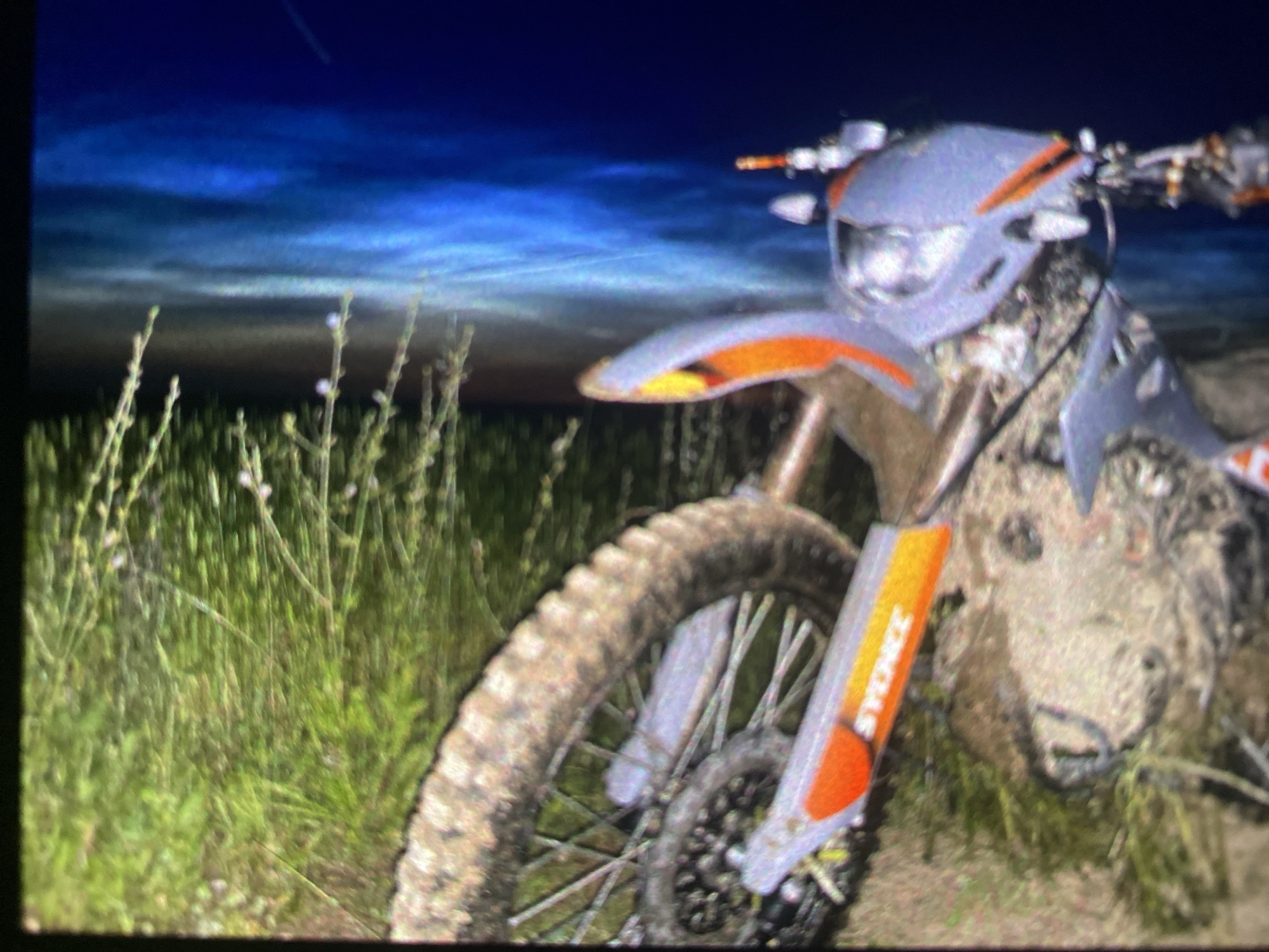 The image shows the front section of a dirt bike positioned amidst tall green grass and weeds, set against a dramatic twilight or nighttime sky. The bike, which features orange and grey bodywork, including the front fender and headlight assembly, is heavily coated in mud, particularly on the front wheel, tire, and lower chassis, indicating recent off-road use. The knobby front tire is prominent on the left side of the foreground. On the right front fork protector, partially obscured text can be seen, appearing to read "SYERGEZ" or a similar word.
The background sky is a deep blue, transitioning to lighter, streaky blue and grey cloud formations near the horizon. These cloud patterns are consistent with twilight or very early morning conditions and could potentially be noctilucent clouds, often observed in high-latitude regions like Saint Petersburg. A faint, thin line, possibly a contrail, is visible in the upper sky. The overall lighting is dim, suggesting either night or the pre-dawn hours. The scene is an outdoor, natural environment, likely a field or off-road trail.