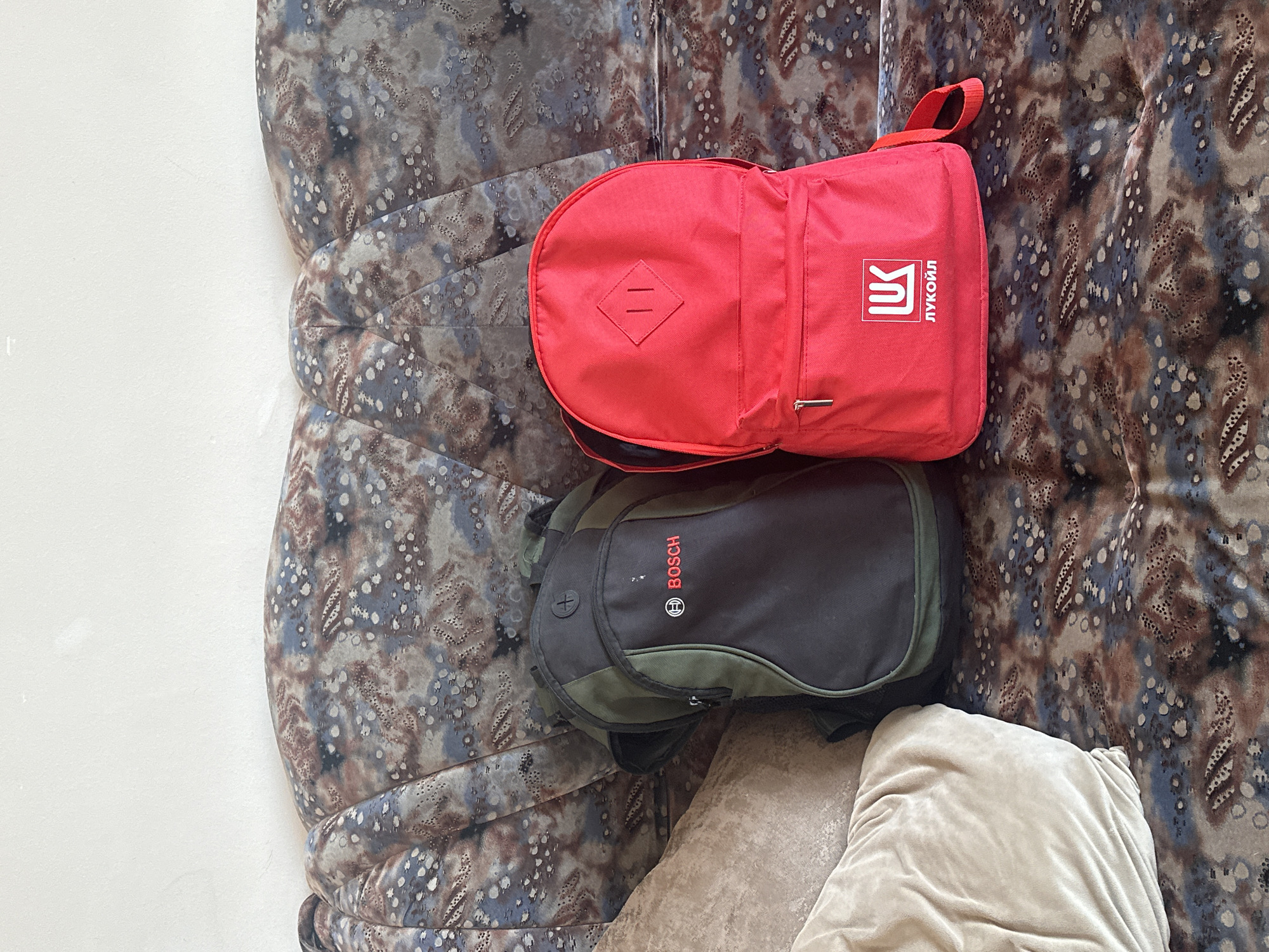 Two backpacks are placed on a patterned sofa. The backpack on the left is green and black and has a "BOSCH" logo. The backpack on the right is red and has a white logo with the word "ЛУКОЙЛ" underneath it. A light brown pillow is to the left of the backpacks. The sofa has a swirling, abstract pattern in shades of brown, blue, and grey, with white speckles. The background is a plain white wall. The image is taken from a high angle, looking down at the backpacks. The location is not specified in the image, and there is no information to suggest it is in Gyumri, Armenia.