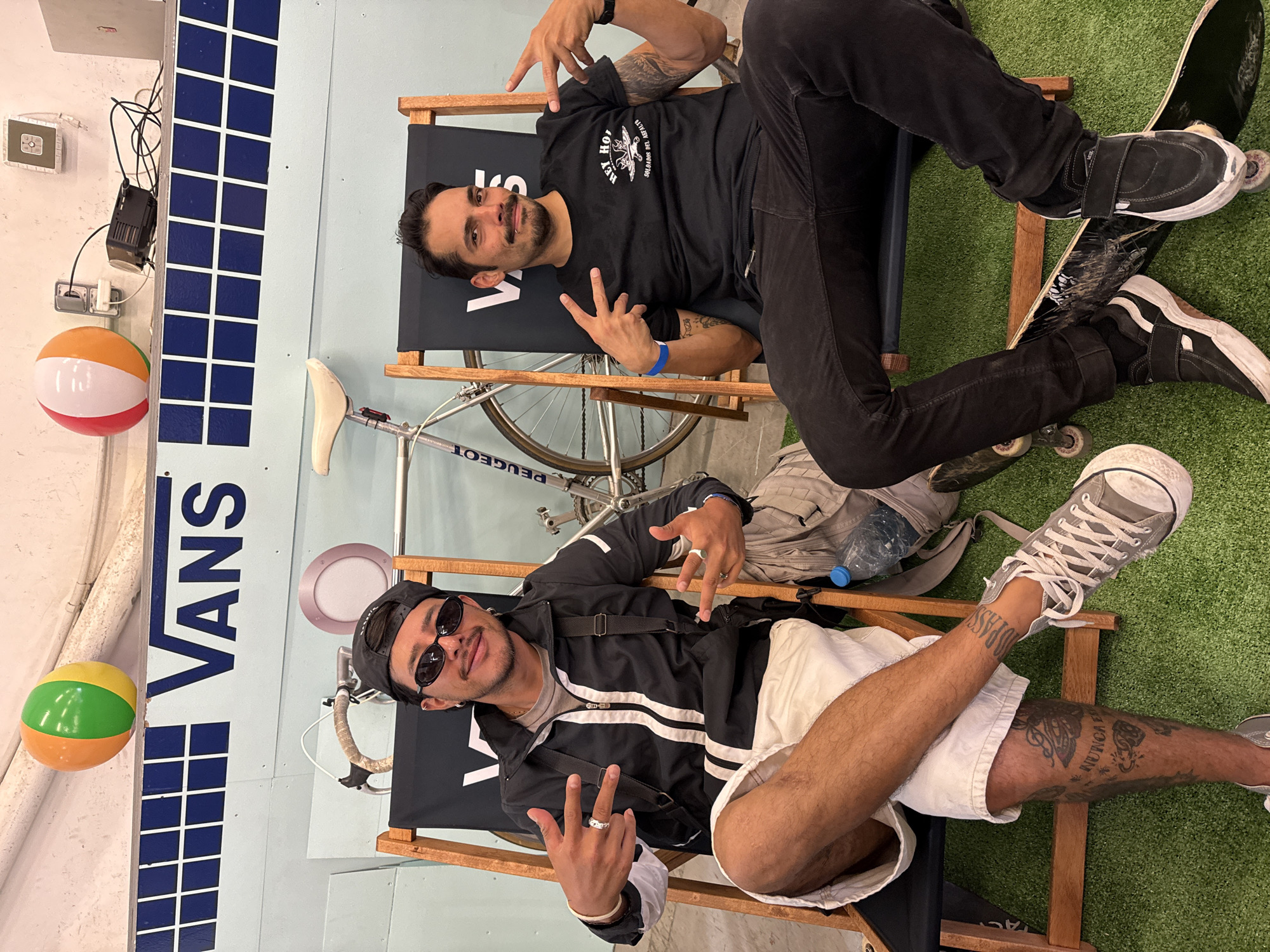 Two men are sitting in beach chairs in front of a backdrop featuring the word "Vans" and a blue and white checkered pattern. A bicycle is positioned behind the chairs, and two beach balls are affixed to the backdrop. The man in the foreground is wearing sunglasses, a black cap, a white-striped black jacket, and white shorts. The man in the background is wearing a black T-shirt and black pants. They are both making hand gestures. Skateboards are on the floor near their feet. The scene appears to be an indoor setup in the city of L'Hospitalet De Llobregat, Spain.