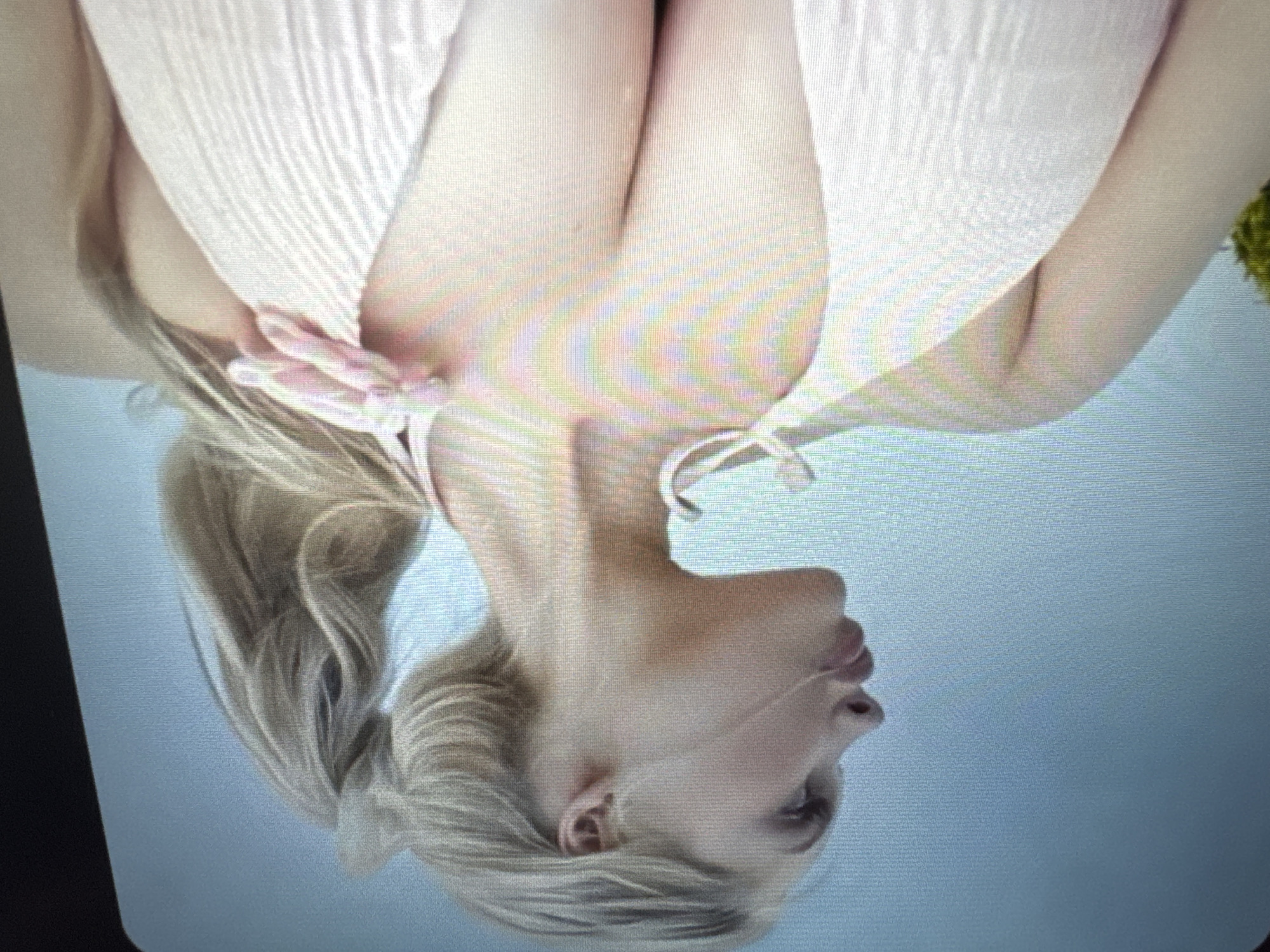 The primary image displays an inverted portrait of a light-skinned person, likely female, featuring blonde hair pulled into a ponytail. The individual's head is positioned at the bottom of the frame, looking towards the right with a slightly open mouth and upward gaze. Their upper torso is visible, clad in a light-colored (possibly white or pale pink) two-piece swimsuit top with discernible tie straps. The background is predominantly a smooth, light blue, suggesting an open outdoor environment such as sky or water, with a small hint of green foliage visible on the far right edge. The perspective is from below, looking up at the subject. The image, along with its provided crops, shows digital artifacts like pixelation and vertical banding, suggesting it may be a photograph of a screen. The crops offer closer details of the swimsuit's ties, the contours of the chest and arms, and the profile of the face and ear. No discernible activity beyond posing for the photograph is evident. There is no visible text. The lighting is soft and even.