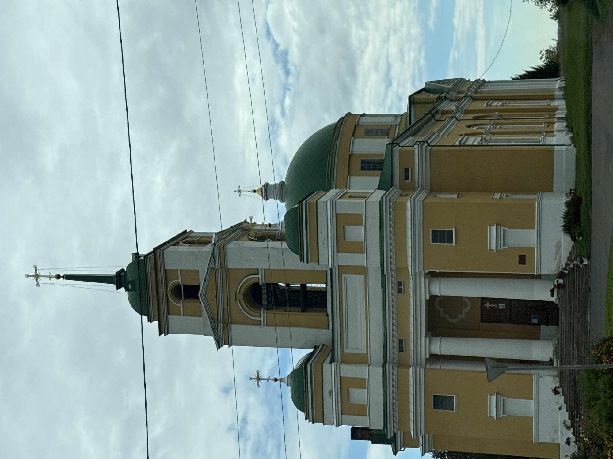 A medium shot captures an Eastern Orthodox church, painted a pale yellow with white trim, under a cloudy sky. The church features a prominent bell tower with a dark green spire topped by a golden cross. Additional green domes with golden crosses are visible on smaller sections of the building. The facade is accented with large white columns flanking the entrance. Steps lead up to a dark wooden door. The church is situated in the village of Mazunino, Russia, with a grassy area in the foreground. Power lines are visible across the top portion of the image.