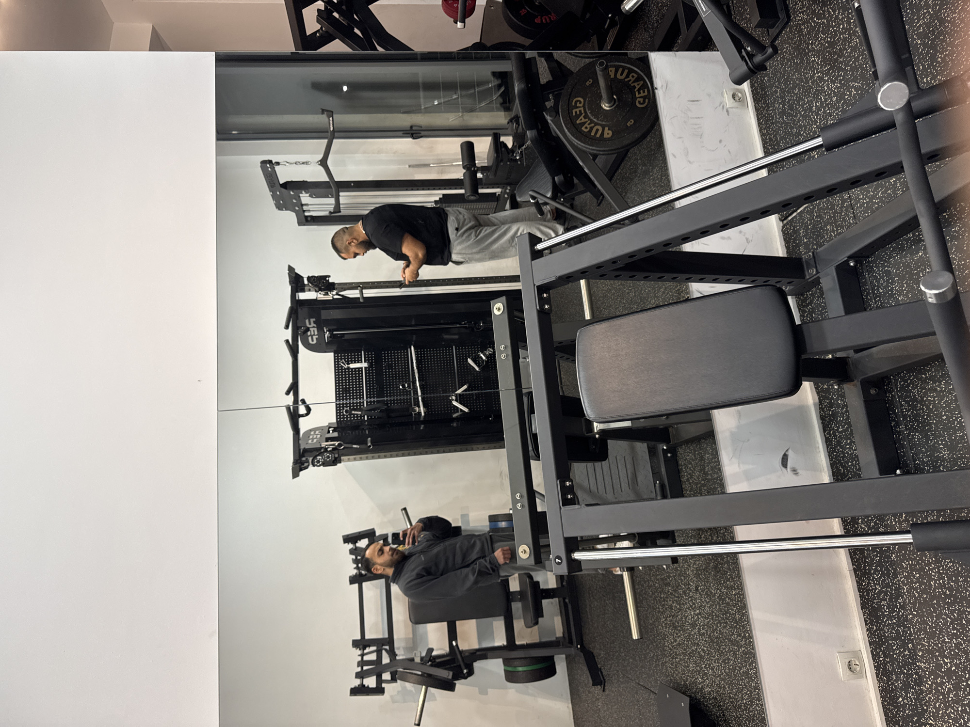 This image captures an interior view of a fitness facility, identified as a gym, located in Plataniás, Greece. The floor is uniformly covered with black, speckled rubber gym flooring. The walls are white, with a prominent mirror covering the lower half, reflecting the interior space.
In the immediate foreground, a black leg press machine, featuring a padded seat and silver support rods, is partially visible. To the left within the mirror's reflection, a male individual with dark hair and a beard is seen wearing a dark grey quarter-zip jacket and grey sweatpants. He holds a smartphone in his right hand, angled towards the mirror, indicating he is either taking a photograph or recording video of himself. Behind him, reflected gym equipment, including a machine with green and black weight plates, is visible.
To the right, standing directly in the gym space in front of the mirrored wall, another male individual with dark, short hair and a beard is present. He is wearing a black short-sleeved t-shirt and light grey sweatpants, holding a smartphone with both hands and looking down at its screen. Behind him and to his left, a black multi-functional cable machine, branded "SFS" in its reflection, is visible. Other black gym equipment, including weight racks and machines with reflected red weight plates, are distributed throughout the background. A white electrical outlet is visible on the wall in the reflection near the floor.