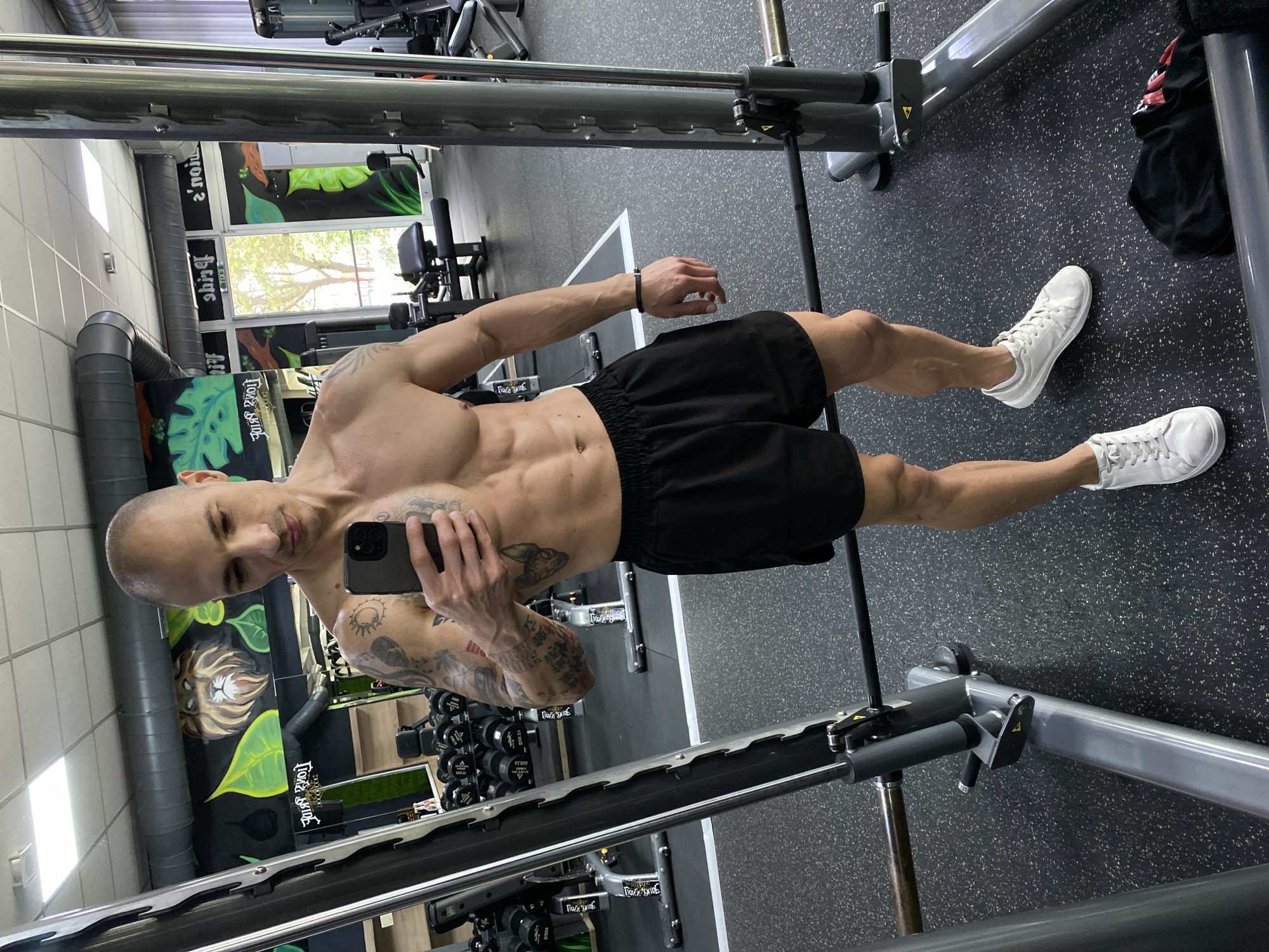 A shirtless man stands in a gym in Pleven, Bulgaria. The man is balding, tanned, and muscular with visible tattoos on his left arm. He wears black shorts and white sneakers. He holds a black smartphone in his right hand, presumably taking a selfie. He appears to be standing in a Smith machine. Dumbbells, weight machines, and muraled walls are visible in the background. The floor is black with a speckled pattern.