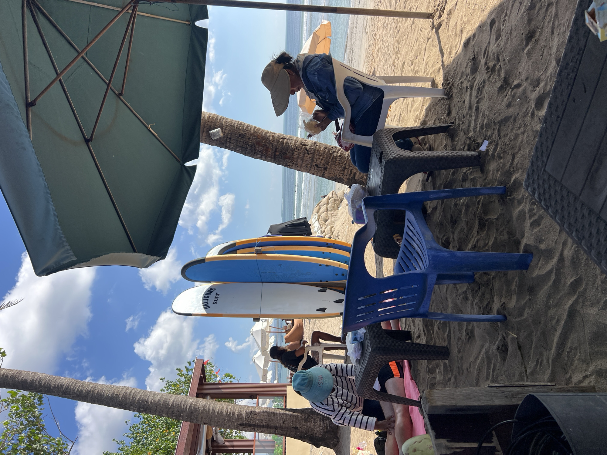 This image depicts a beach scene in Kuta, Indonesia, likely on a sunny day with a clear blue sky and scattered clouds. In the foreground, a person wearing a striped long-sleeved shirt and a blue hat is giving a massage to another person lying on a pink mat on the sand. A dark woven table sits in front of them. To the right, a woman wearing a denim jacket and a wide-brimmed hat is sitting in a white plastic chair under a large green umbrella, holding what appears to be a piece of fruit or snack. In the background, several surfboards stand upright. Beyond the beach, the ocean is visible with gentle waves and a few distant surfers. Palm trees and a beachside structure are also present, contributing to the tropical atmosphere of Kuta. The sand is a light brown, and shadows indicate the sun is high in the sky.