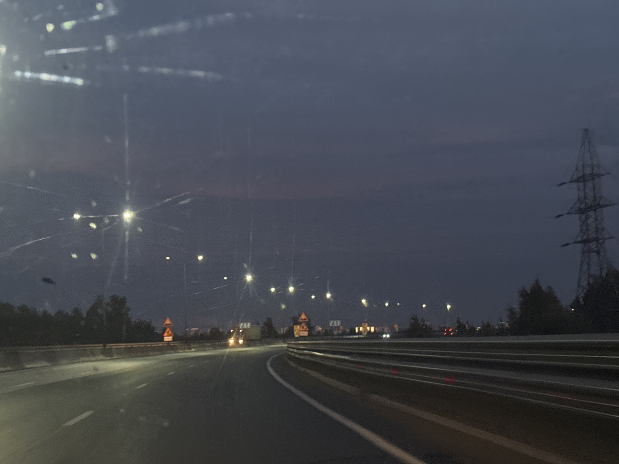 The image depicts a nighttime scene on a highway, likely on the outskirts of Perm, Russia. The sky is dark and overcast, with a faint glow suggesting twilight or early evening. The highway curves to the right, with multiple lanes of asphalt. A metal guardrail runs along the right side of the road, reflecting red and white lights. Streetlights are visible along the left side of the highway, casting pools of light onto the road. In the distance, trees and buildings are silhouetted against the sky, and a tall electricity pylon stands on the right. There are no people or vehicles clearly visible in the foreground, but distant headlights suggest other traffic. The overall impression is one of a typical evening commute or travel on a major road.