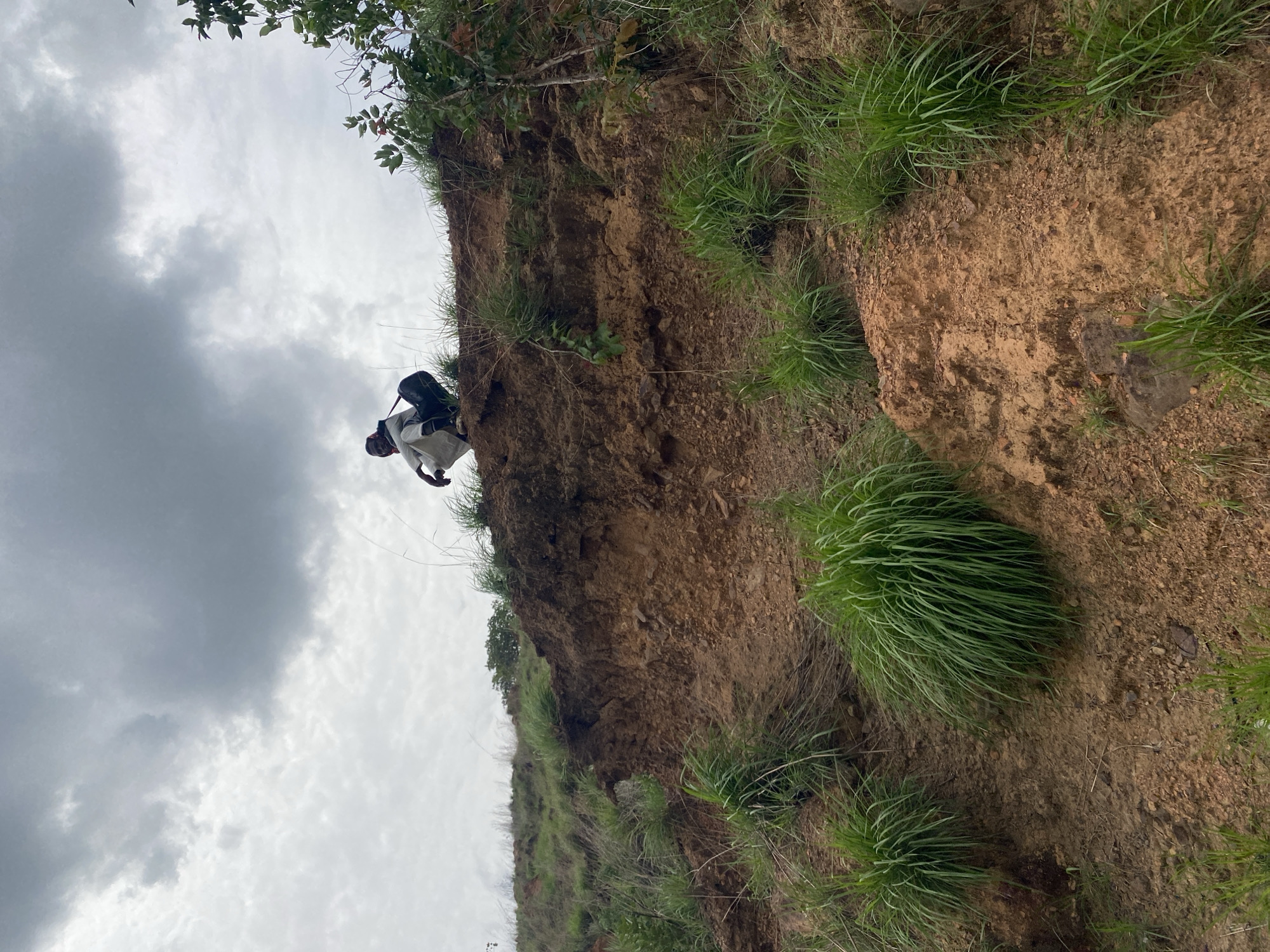 The image displays a person navigating a steep, earthy slope under an overcast sky. The individual, seen from a low angle, appears to be a male wearing a light-colored (possibly white or light grey) long-sleeved top and dark trousers, with a dark backpack or container strapped to their back. They are positioned on the brown soil and rock face, which is interspersed with clumps of bright green grass and some sparse leafy plants. The person's body language suggests they are carefully moving along or climbing the incline. Above, the sky is filled with grey, heavy clouds, indicating an overcast day. The terrain is rugged and natural, characteristic of an undeveloped, rural, or hilly area. No text is visible. The location context provided is Amaigbo, Nigeria.