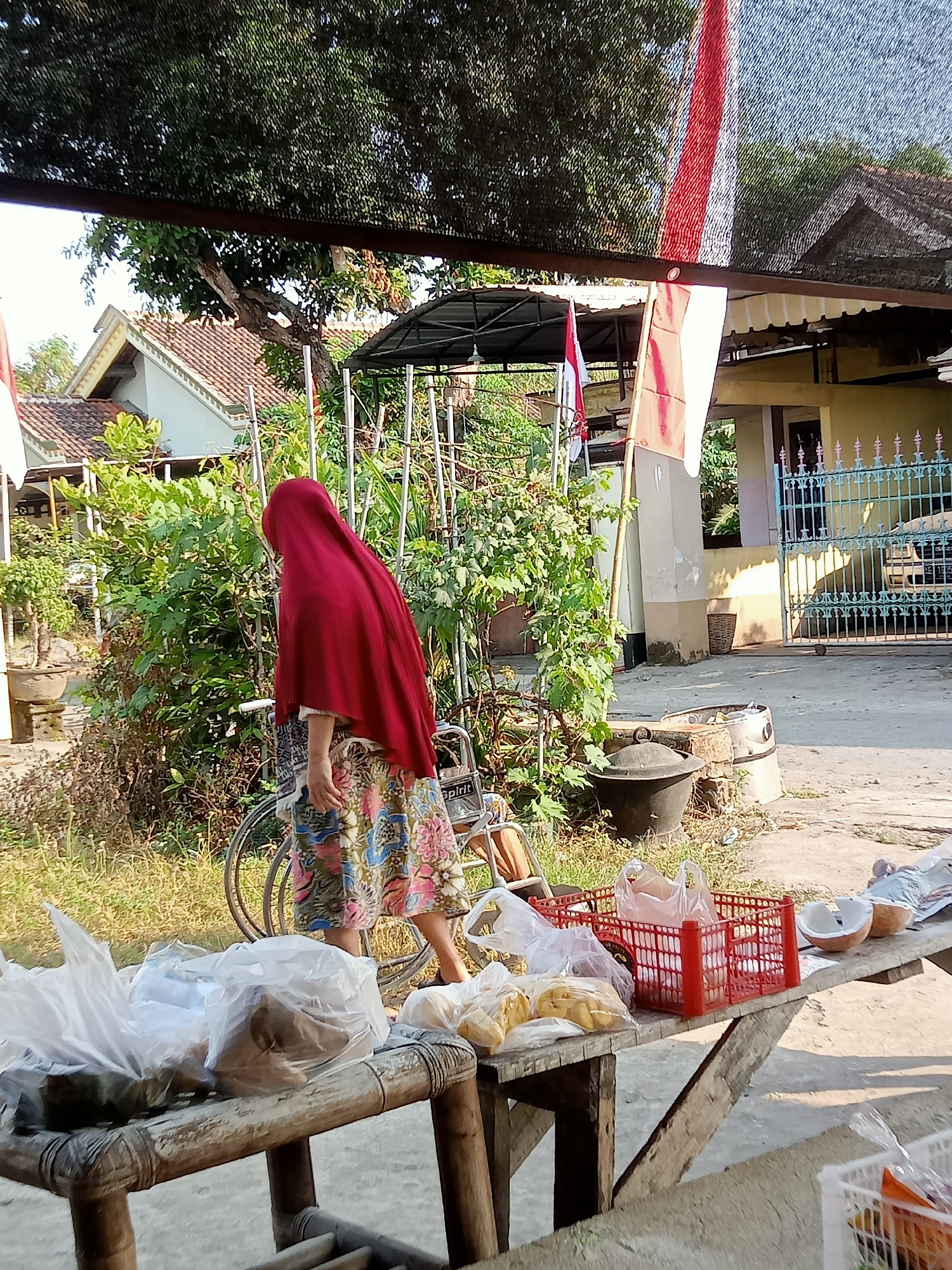 A woman wearing a red hijab and a floral skirt stands next to a bicycle, facing away from the camera. She is positioned on a dirt path in what appears to be a residential area. In the foreground, a wooden table displays various items, including plastic bags filled with goods, a red plastic crate, and two halved coconuts. Behind the woman, a lush green bush grows. The background shows houses with tiled roofs, a blue metal fence, and trees. A flag with red and white stripes is visible hanging from a pole. The scene is illuminated by natural sunlight, casting shadows. The overall impression is that of a street-side market or a household setup in Simo, Indonesia.