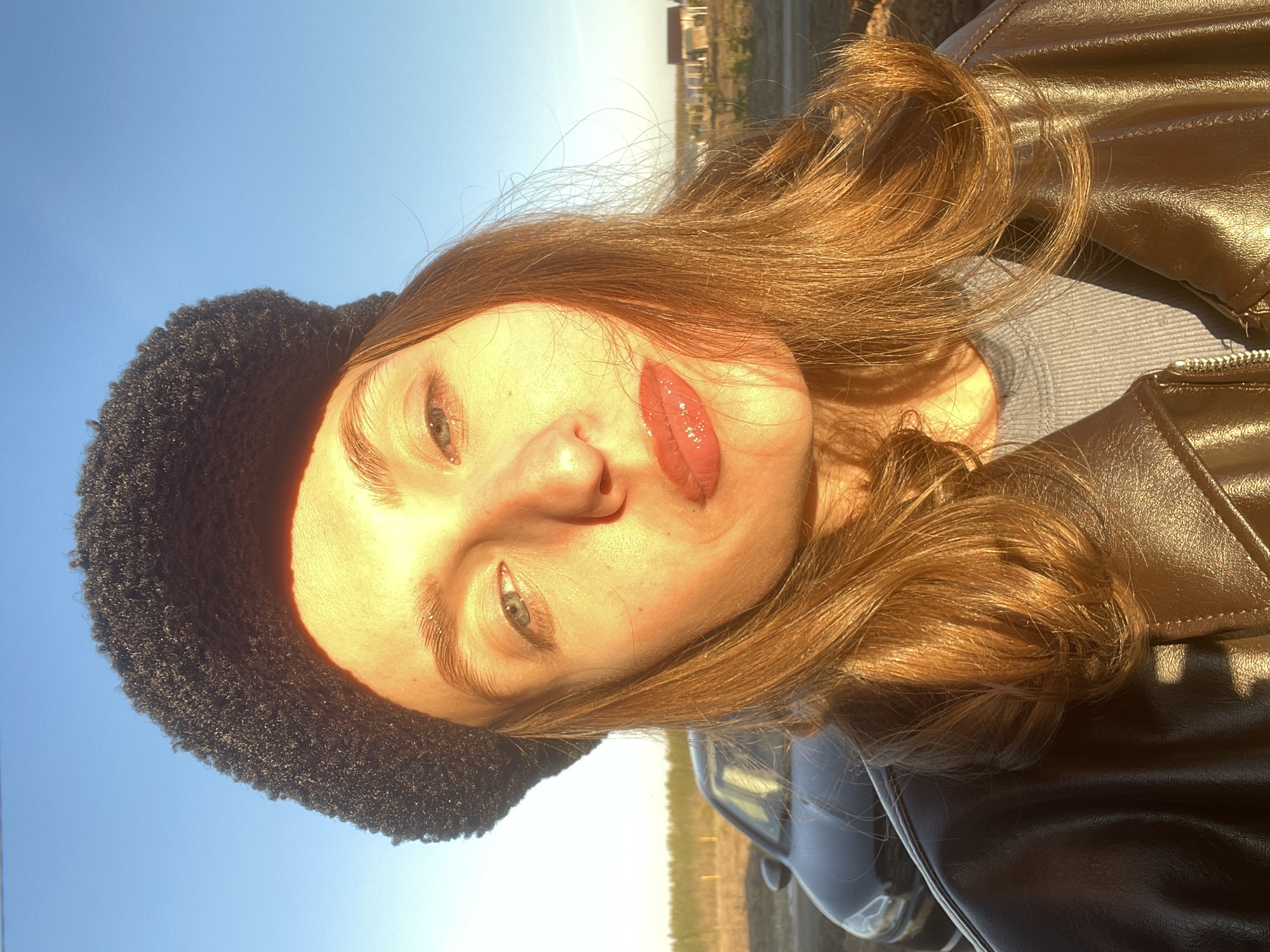 A brightly lit, close-up, and slightly tilted portrait captures a young individual outdoors. The subject, likely female, has fair skin, light-colored eyes (appearing greenish-blue), and long, wavy reddish-brown hair. She wears a dark, textured hat, possibly faux fur, on the left side of her head, and her lips are adorned with a glossy, reddish-pink product. She gazes directly at the camera with a neutral or gently serene expression.
She is dressed in a dark brown or bronze-colored faux leather jacket, visible on her right side, over a light grey or blue ribbed top. The scene is bathed in warm, golden light, strongly indicating either sunrise or sunset, casting a radiant glow on her face and hair. Above her, the sky is a clear, vibrant blue.
The blurred background reveals an expansive outdoor landscape, with what appears to be fields or undeveloped land stretching to a low horizon. Distant structures, possibly houses or buildings, are faintly visible along the horizon line. A partial view of a vehicle, likely a car, is also present in the lower right portion of the frame. The overall setting suggests an open, natural environment, consistent with the provided context of Podshivalovo, Russia. The individual appears to be taking a selfie. No visible text is present in the image.