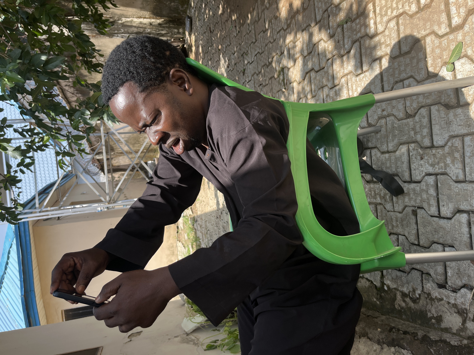 A man is outdoors, with his phone in hand. He is wearing a black, long-sleeved garment. He is leaning forward and looking intently at his phone. A bright green object, possibly part of a chair or a walker, is behind his neck and shoulders. The ground is paved with interlocking concrete bricks, and there is a shadow cast on the ground, indicating it is daytime. Sunlight glints off the man's forehead. He appears focused on his phone, with his mouth slightly open. To the left of the frame, there are some green leaves and what appears to be the roof of a building with blue trim. The scene suggests a casual, everyday moment.